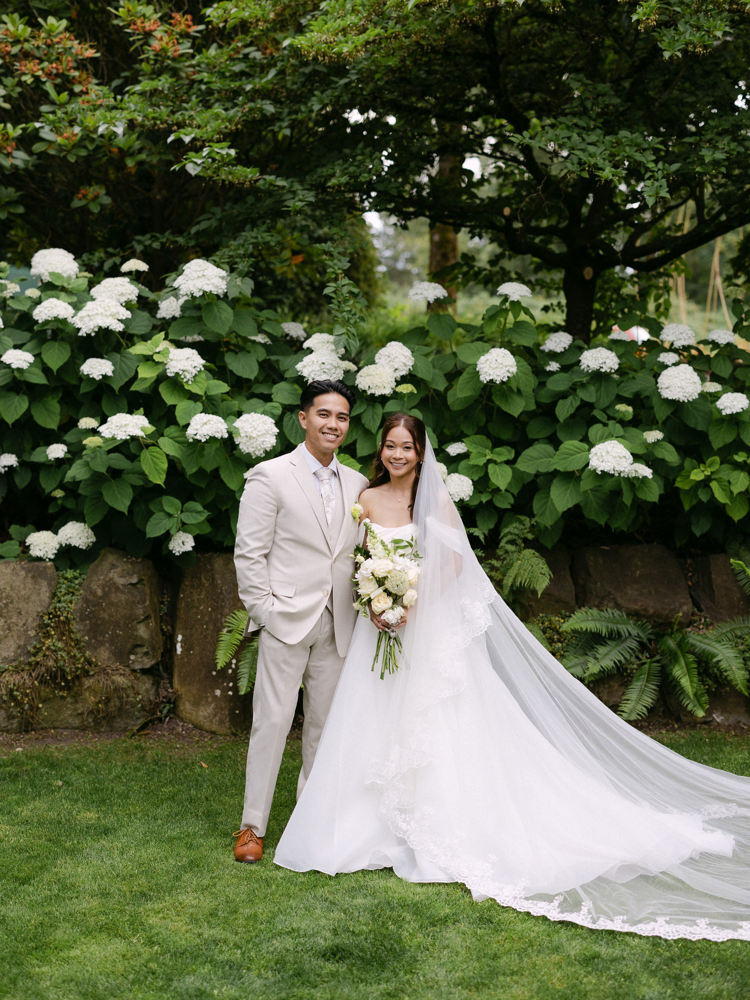 Bride and groom posing before lush white hydrangeas, bride in strapless gown with cathedral veil.