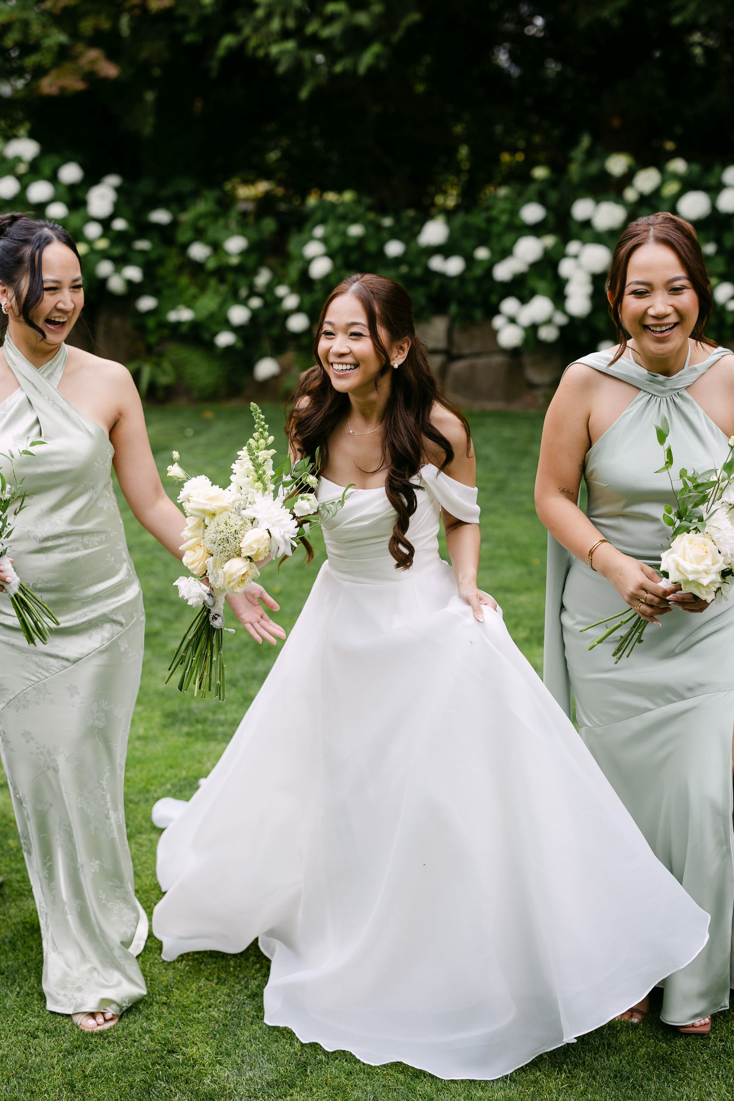 Bride in white off-shoulder gown walks with two bridesmaids in sage green satin dresses, holding white bouquets outdoors.