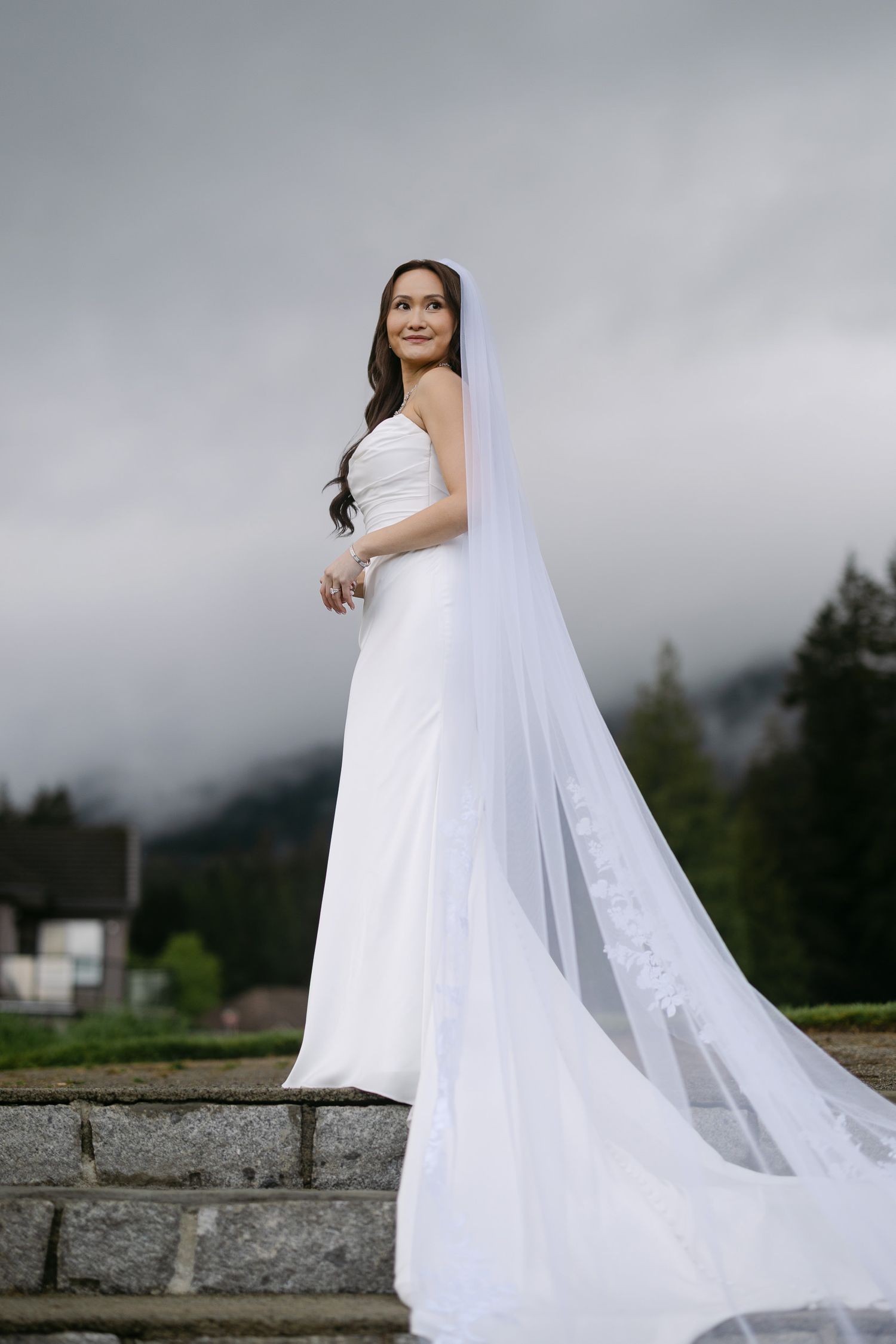 Bride in white gown with lace-trimmed cathedral veil stands on stone steps against cloudy mountain backdrop.