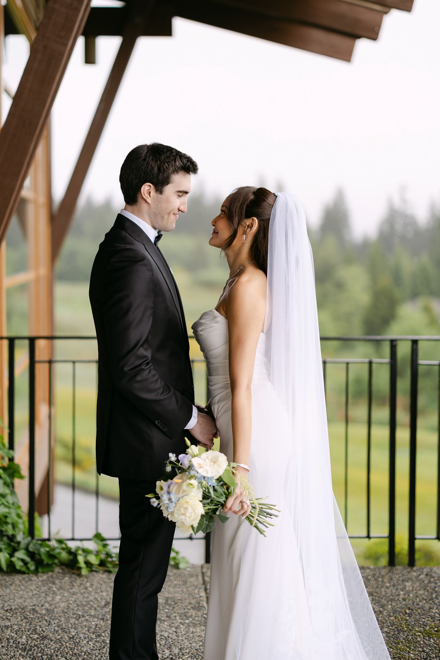 Bride and groom share a romantic moment on an outdoor balcony, she holds white bouquet, green landscape behind them.