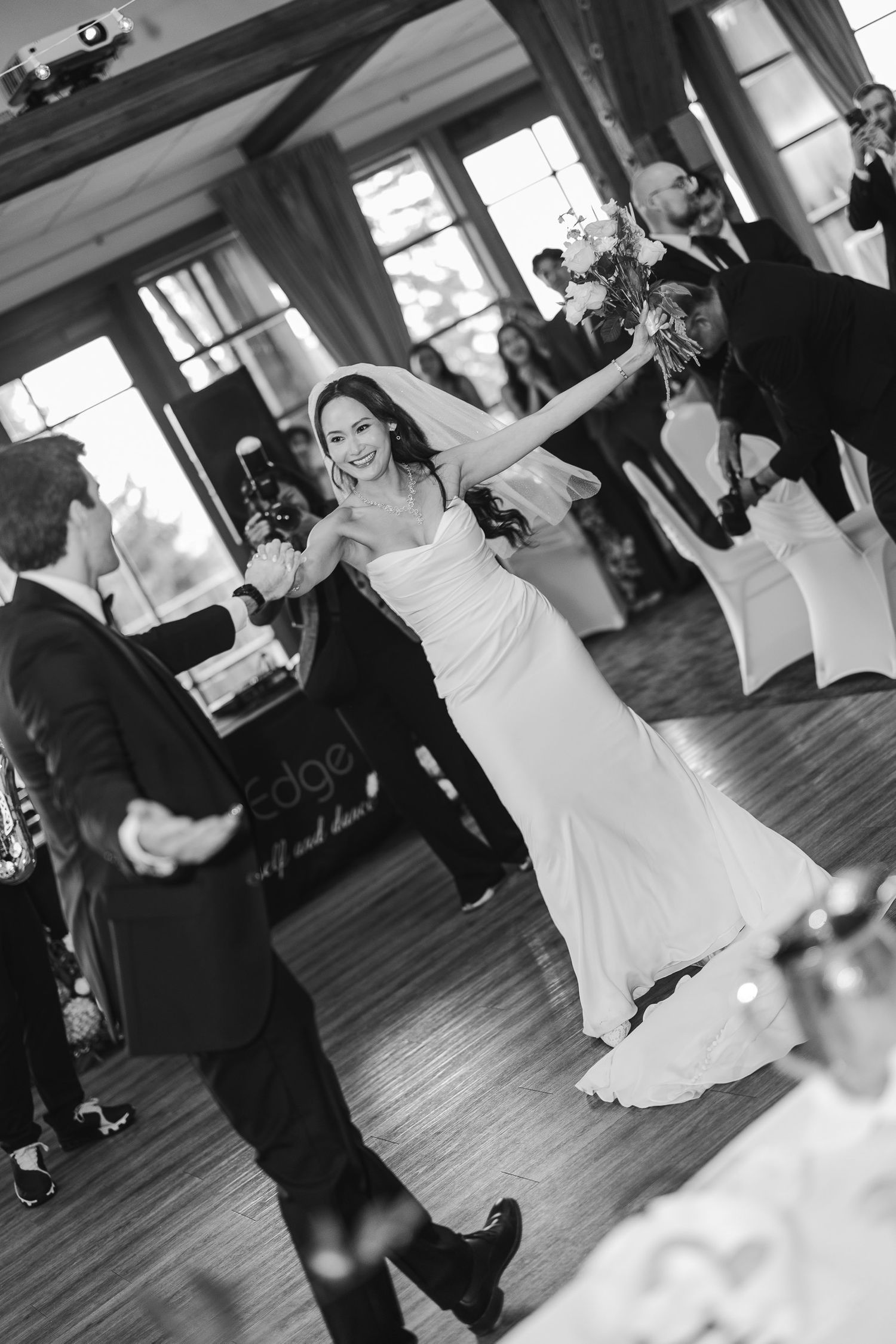 Black and white photo of a joyful bride in a white gown dancing at her wedding reception holding a bouquet.