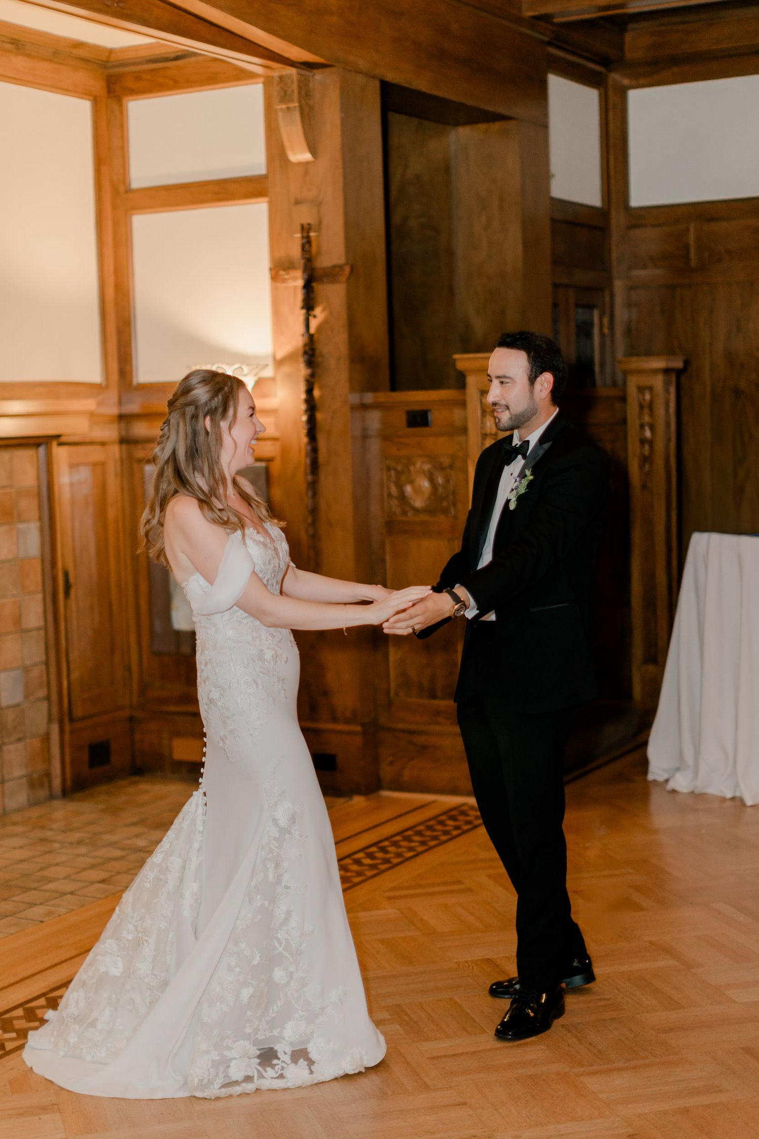 A couple shares their first dance at their wedding reception in an elegant wood-paneled room with hardwood floors.