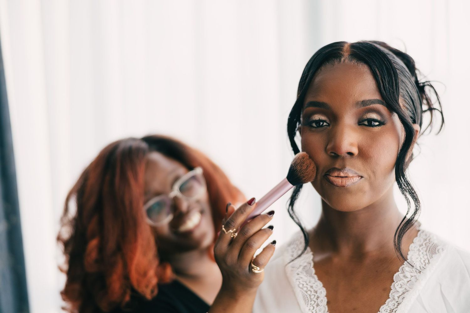 A makeup artist applies lipstick to a bride during wedding preparations while smiling in the background.