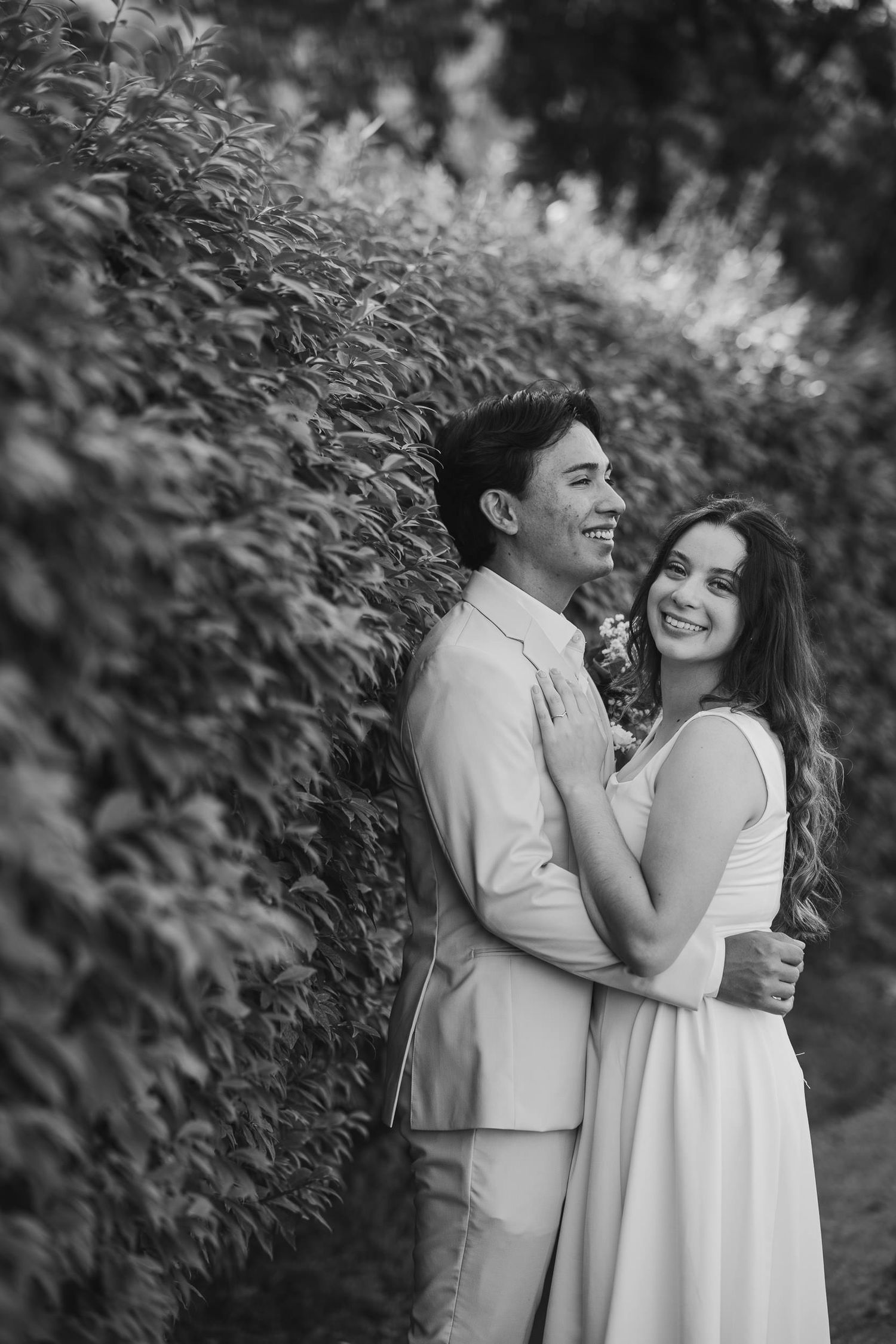 A romantic black and white portrait of a couple embracing and smiling together against a garden hedge background.