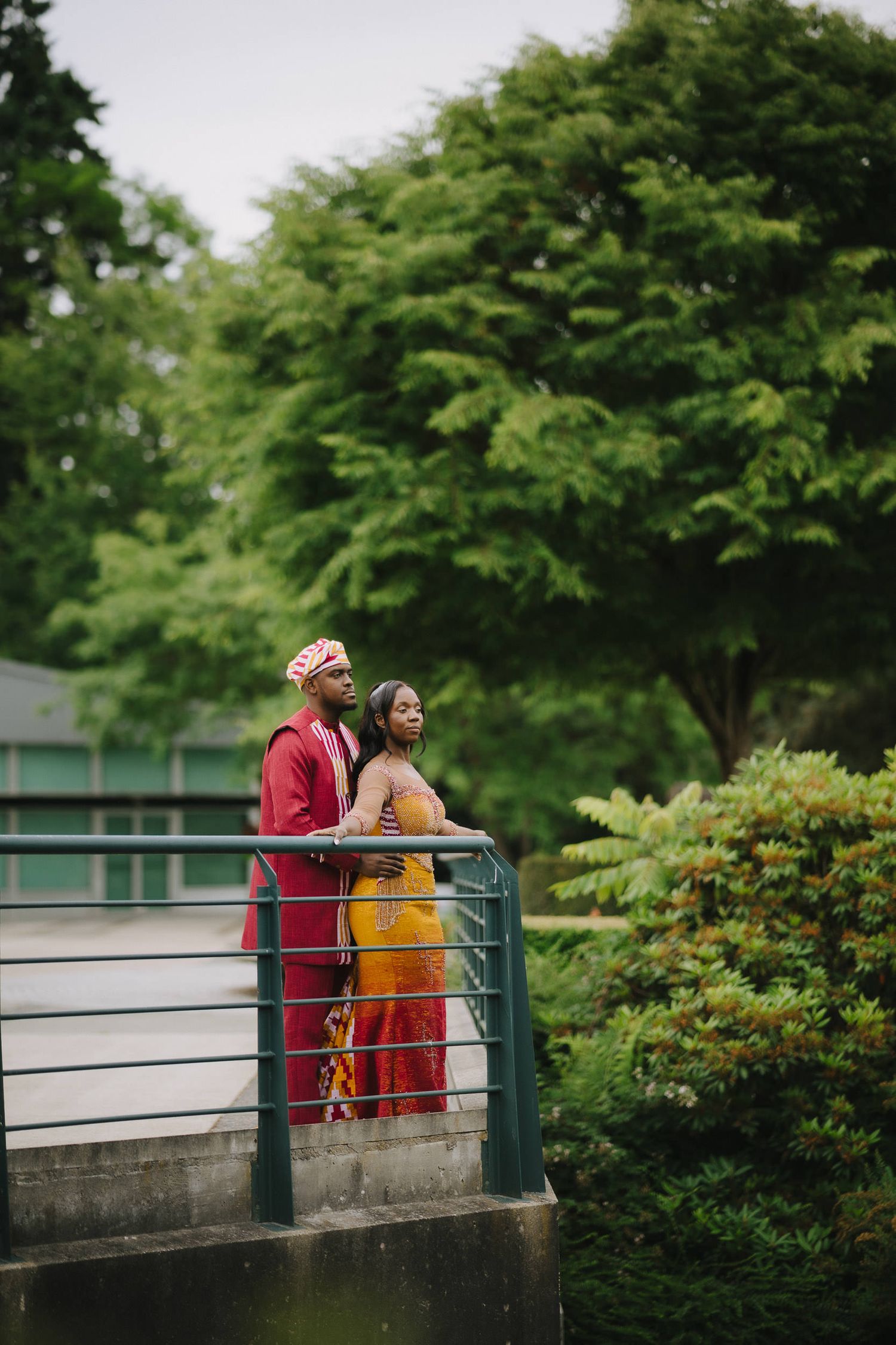 A couple in red and yellow outfits stand together on a bridge railing surrounded by lush green trees.