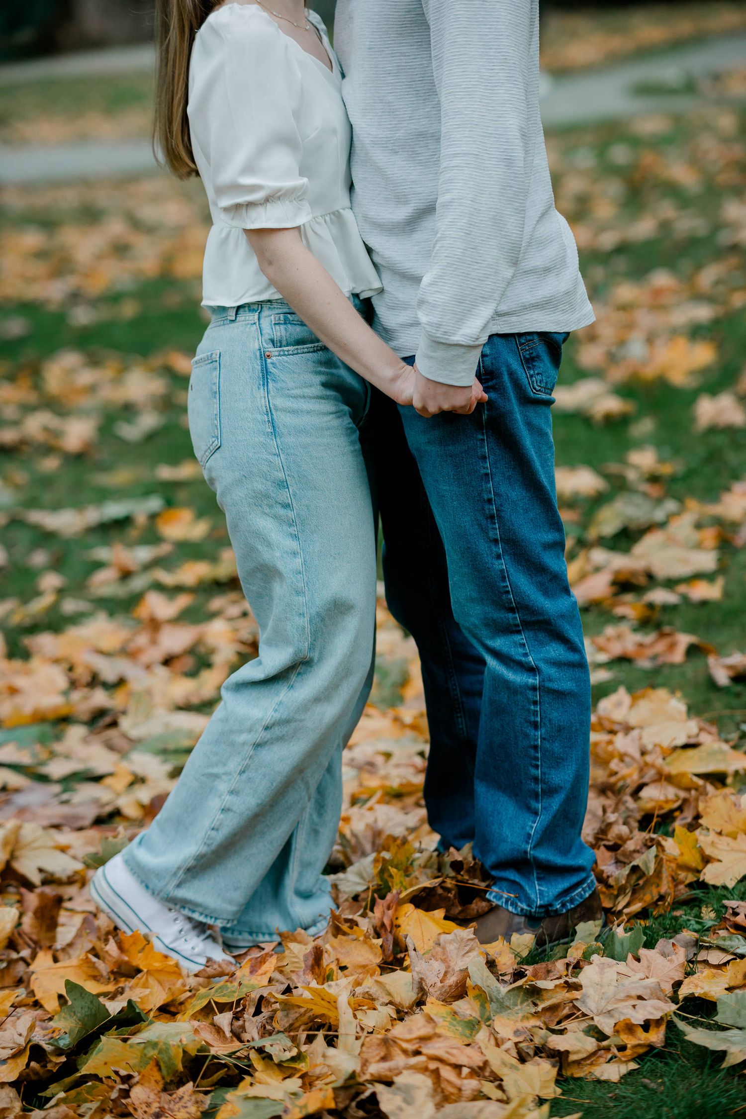 Close-up view of two people standing together in jeans on autumn leaves covering the ground.