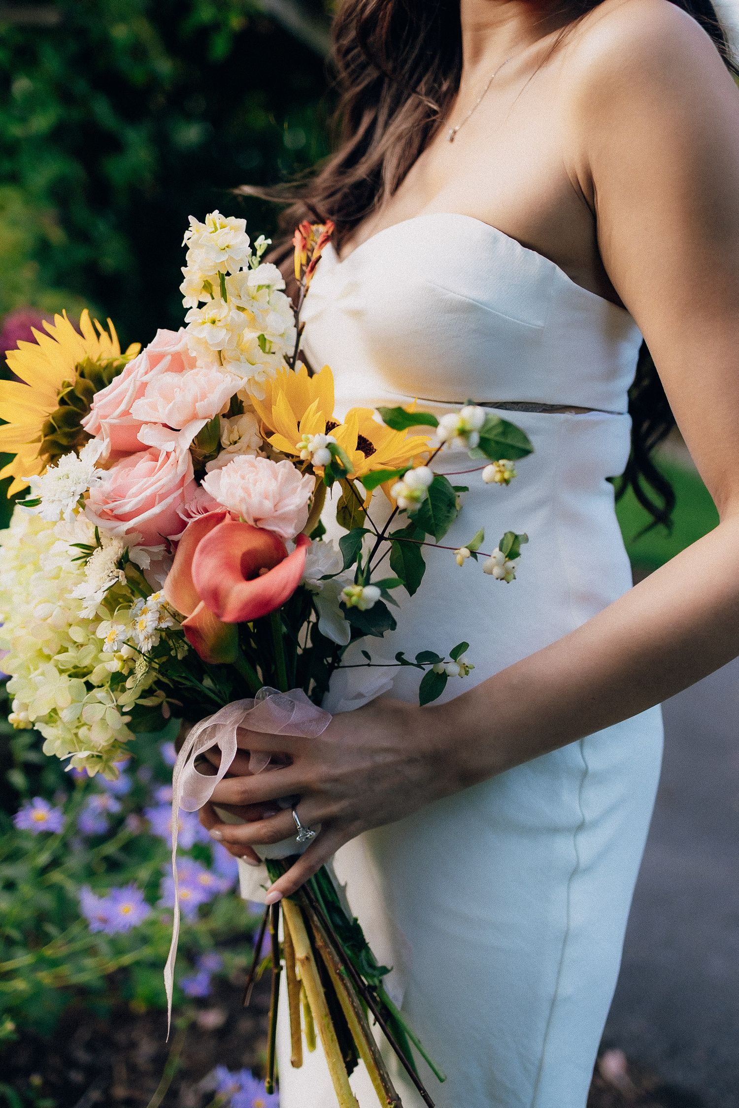 Wedding bouquet with sunflowers, roses and hydrangeas held against a white bridal gown in natural outdoor lighting.