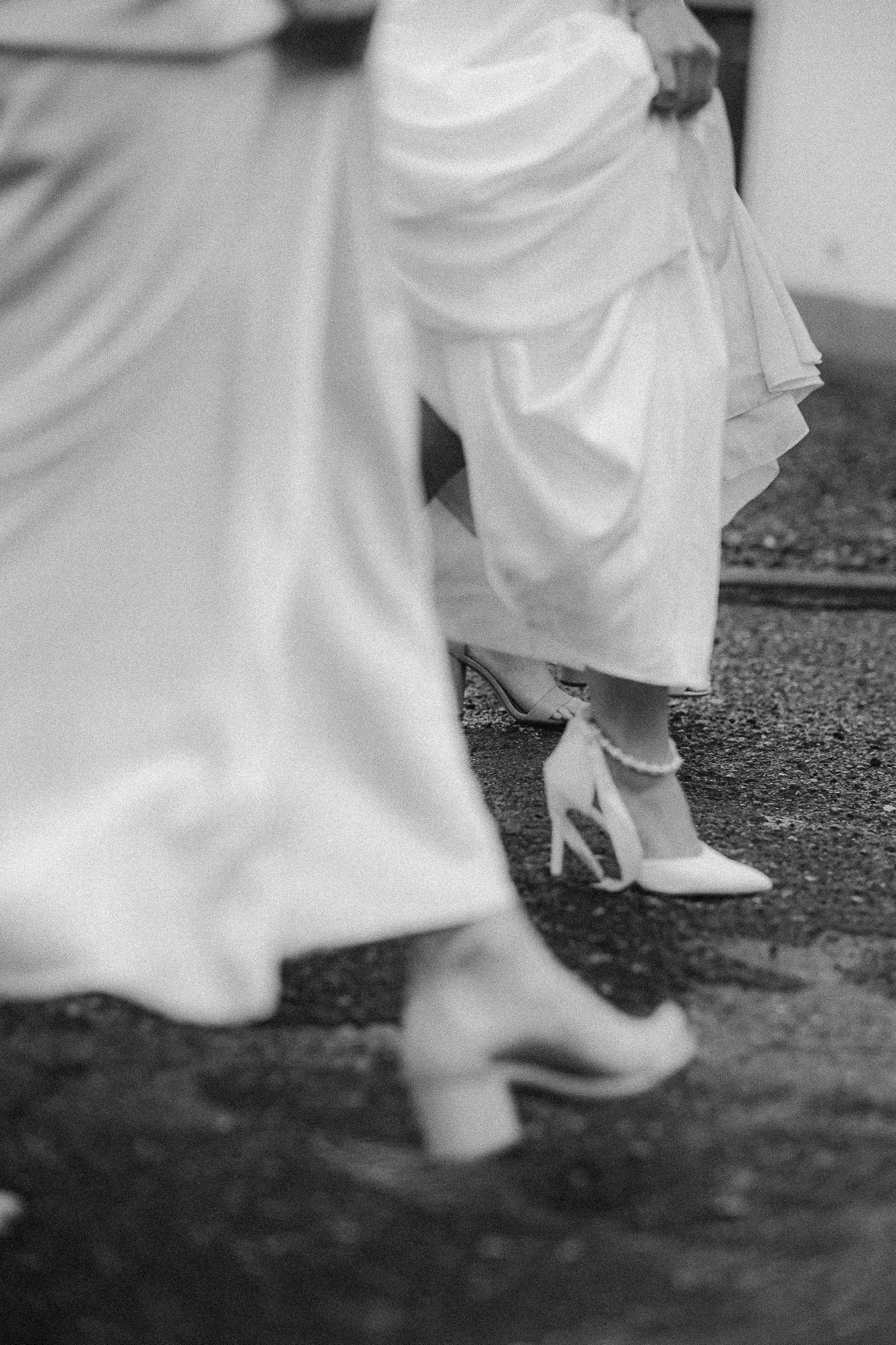 Close-up view of wedding party members' feet and dress hems as they walk on wet pavement.