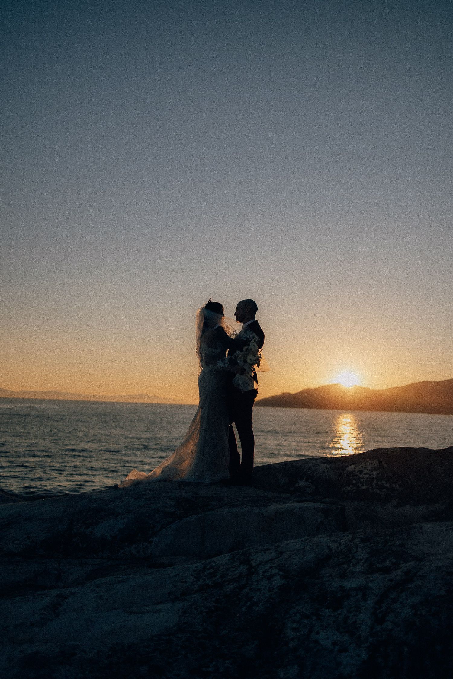 A silhouetted couple embraces on rocky coastal cliffs during a dramatic sunset over the water.