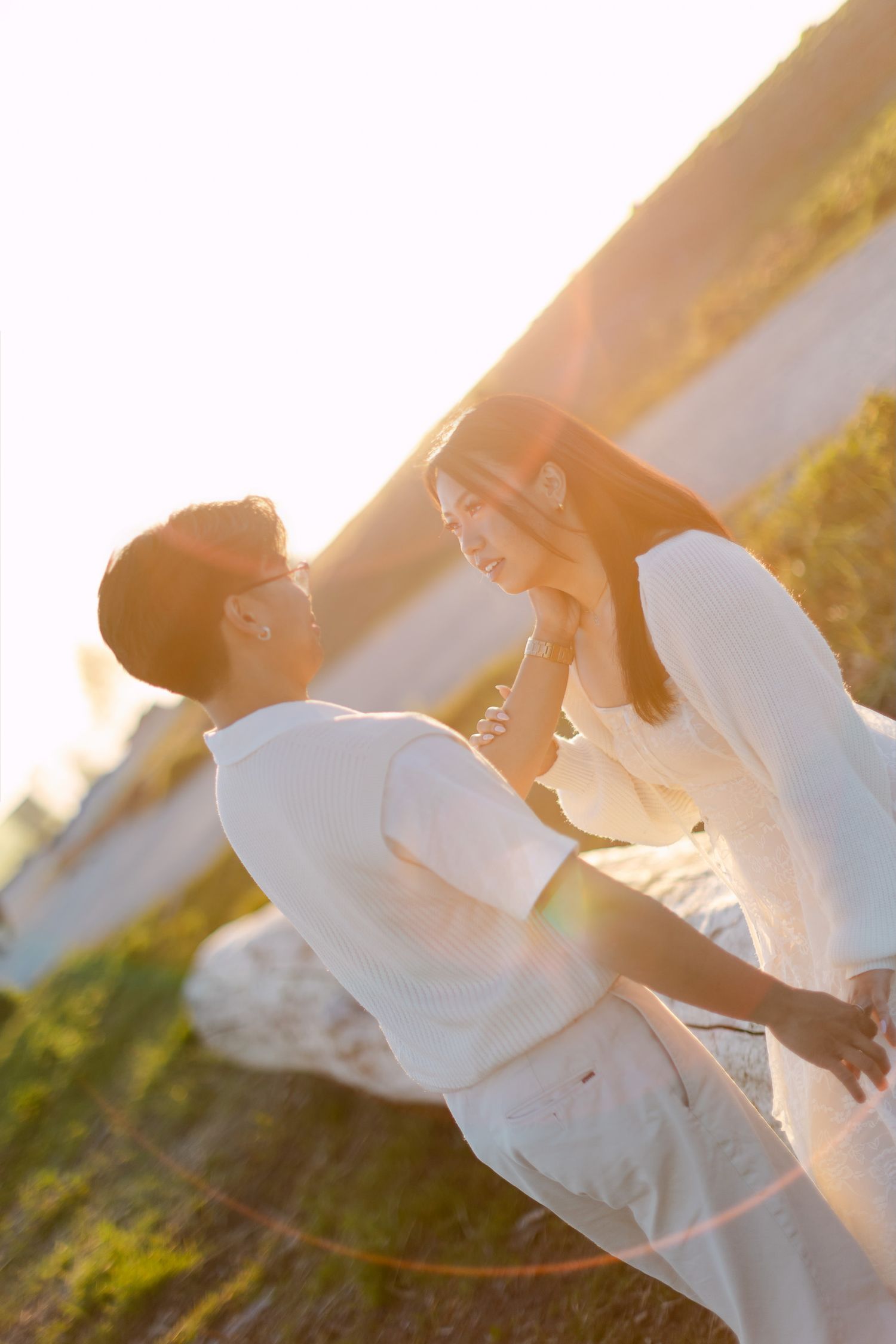 A couple in white clothing shares a tender moment outdoors at golden hour, with warm sunlight flaring behind them.