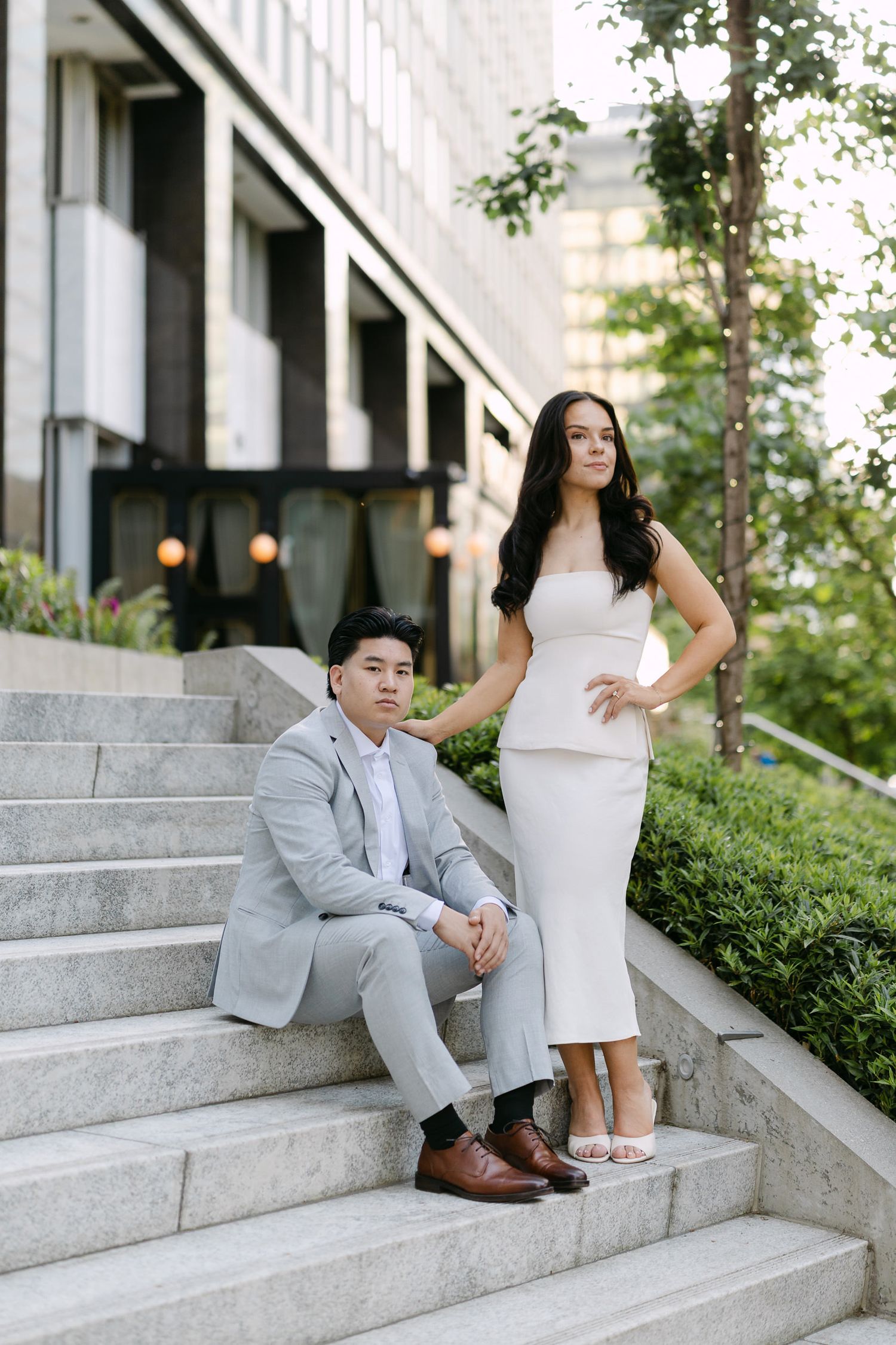 Woman in a white dress stands beside a seated man in a gray suit on city steps lined with trees.