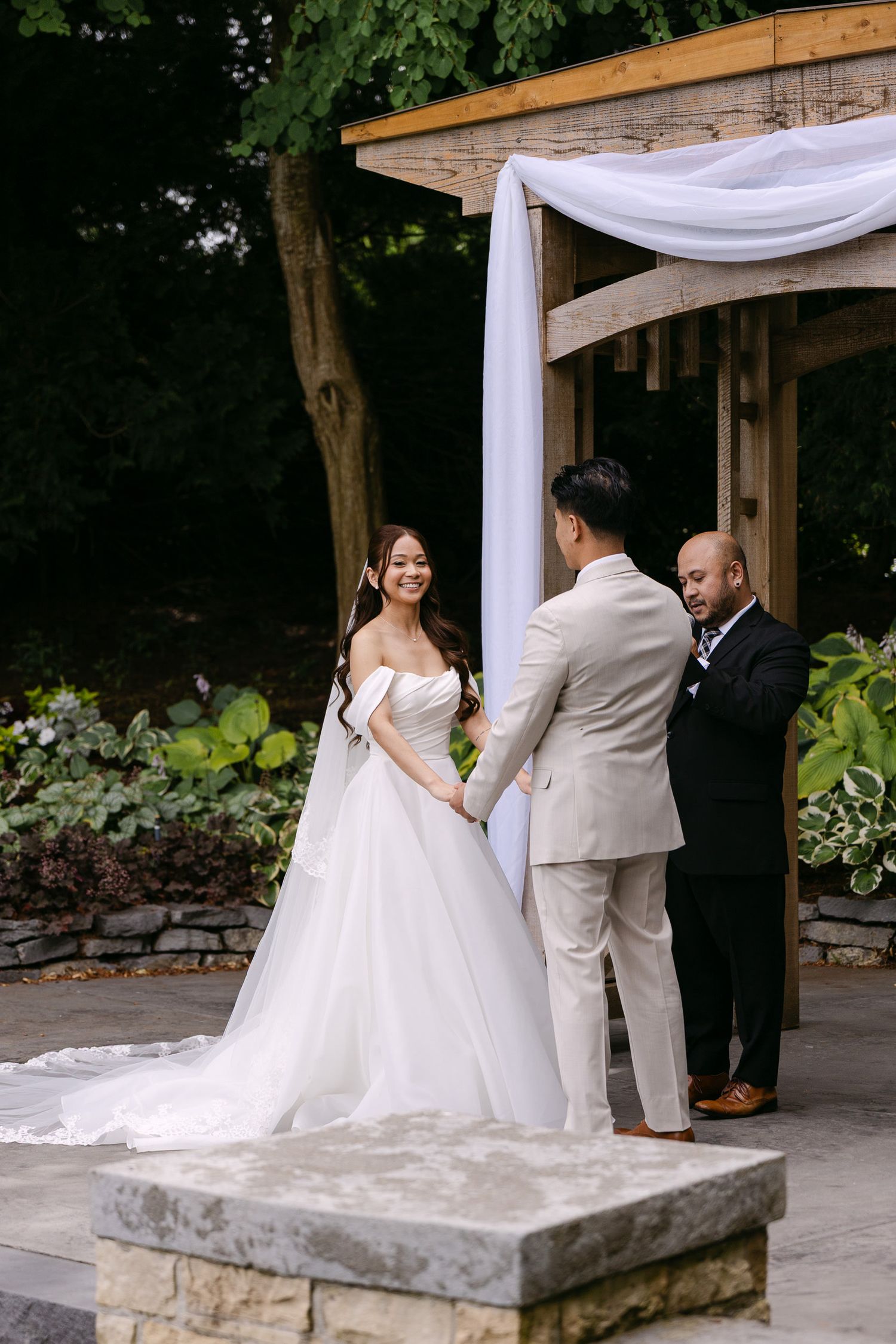 Bride and groom exchange vows outdoors under a white draped wooden arch surrounded by lush greenery.