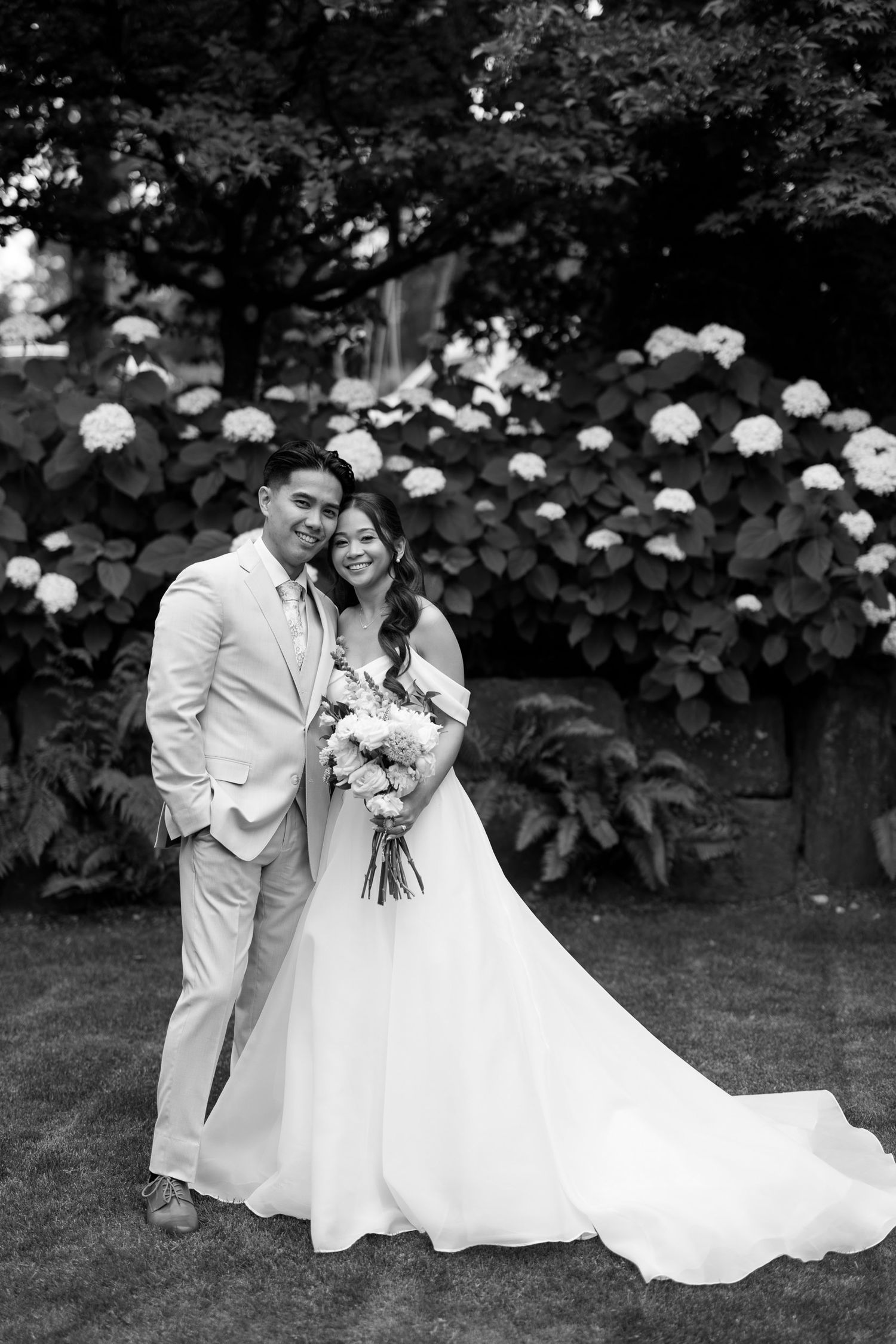 Black and white wedding portrait of a smiling couple posing before blooming hydrangea bushes, bride holding a floral bouquet.