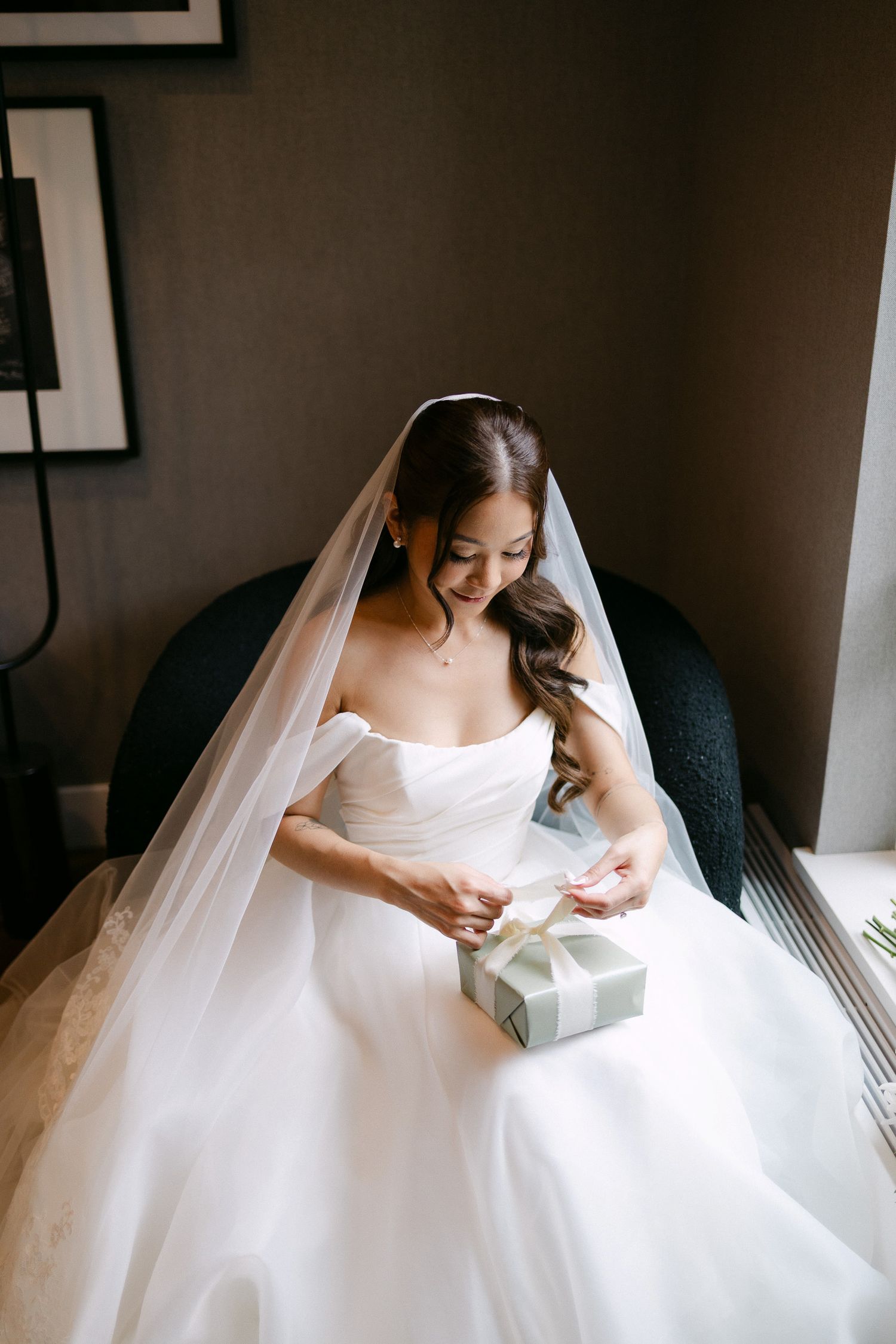 Bride in white gown and veil sits opening a gift box on her wedding day.