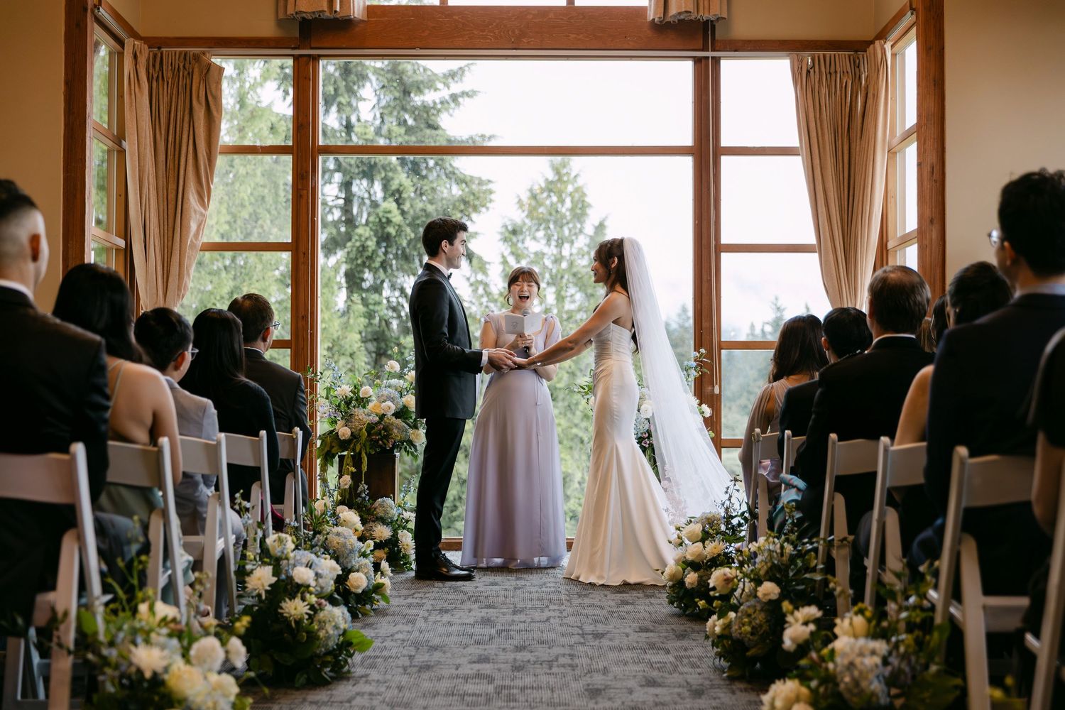 Couple exchanges vows at elegant indoor wedding ceremony with floral aisle and large windows.