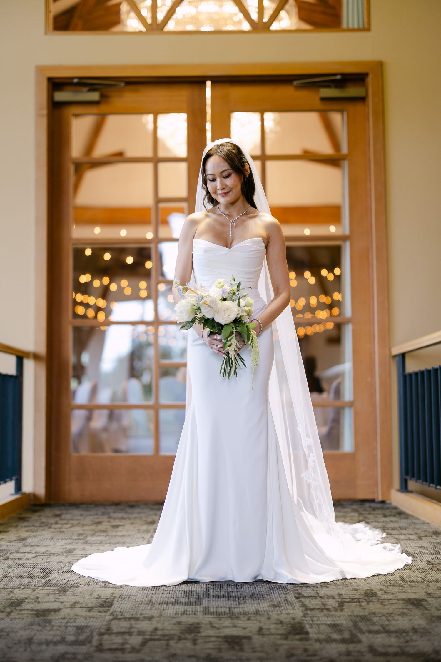 Elegant bride in strapless white gown with long train stands before warm lit doors.