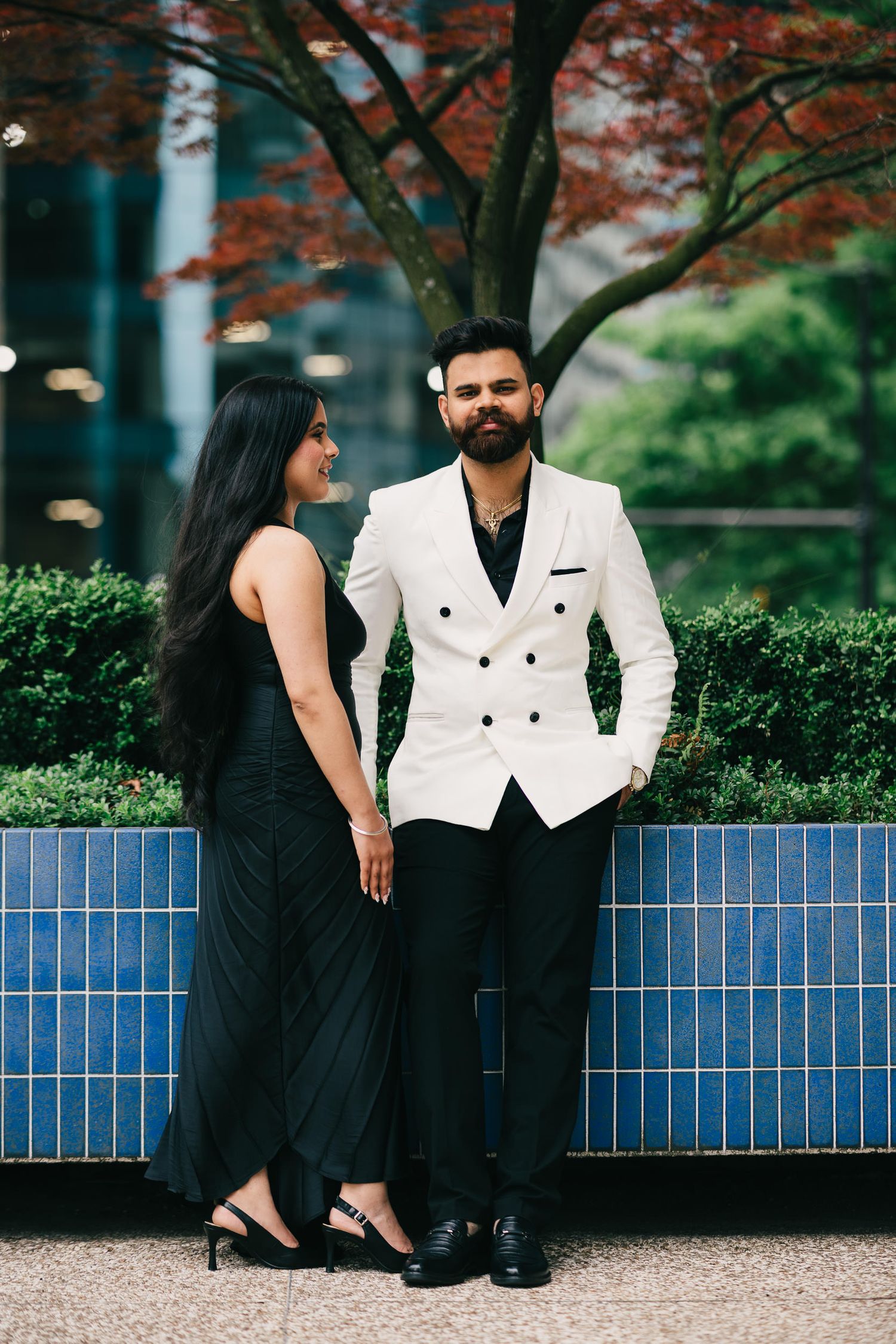 A couple in formal attire poses for romantic engagement photos on a bench under autumn trees with blue tiles behind them.