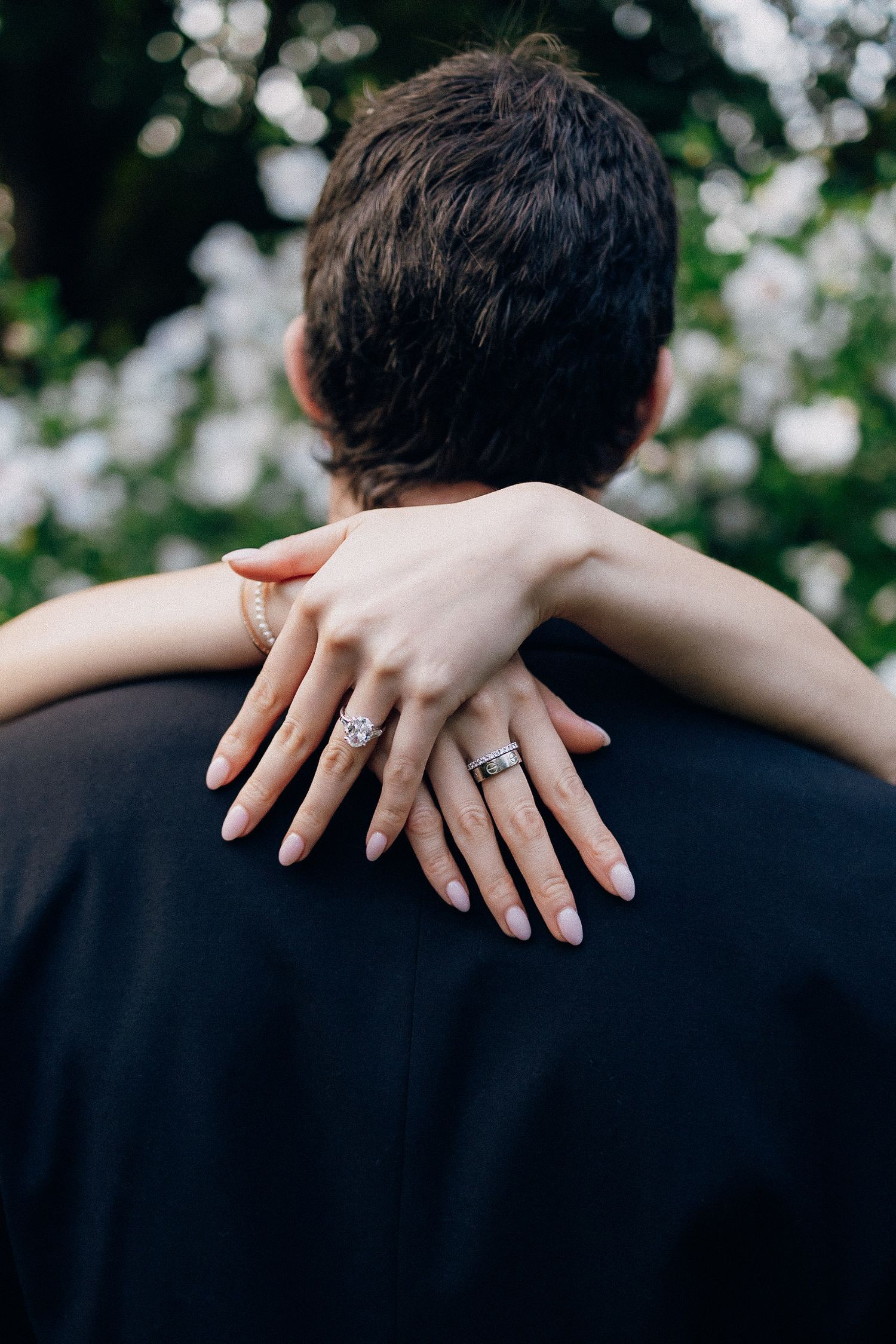 Close-up sequence of hands with engagement ring embracing shoulders against blurred floral background.