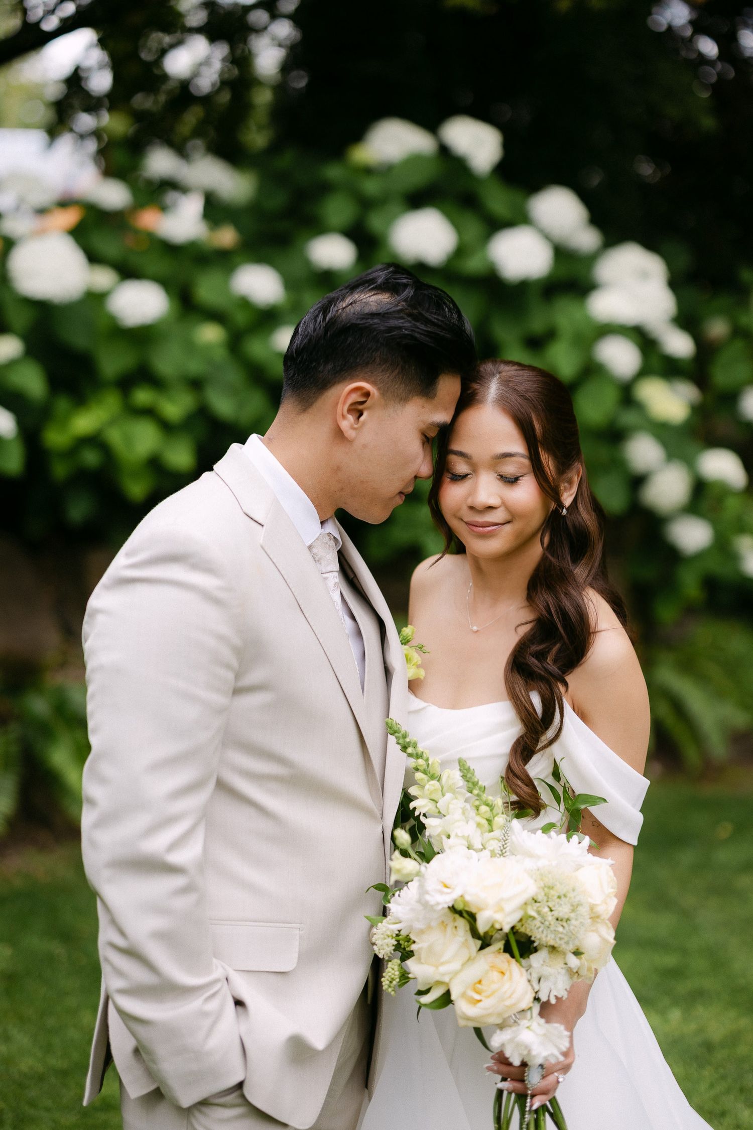 Bride and groom share a tender moment in a garden, surrounded by white hydrangeas, as she holds a lush white bouquet.