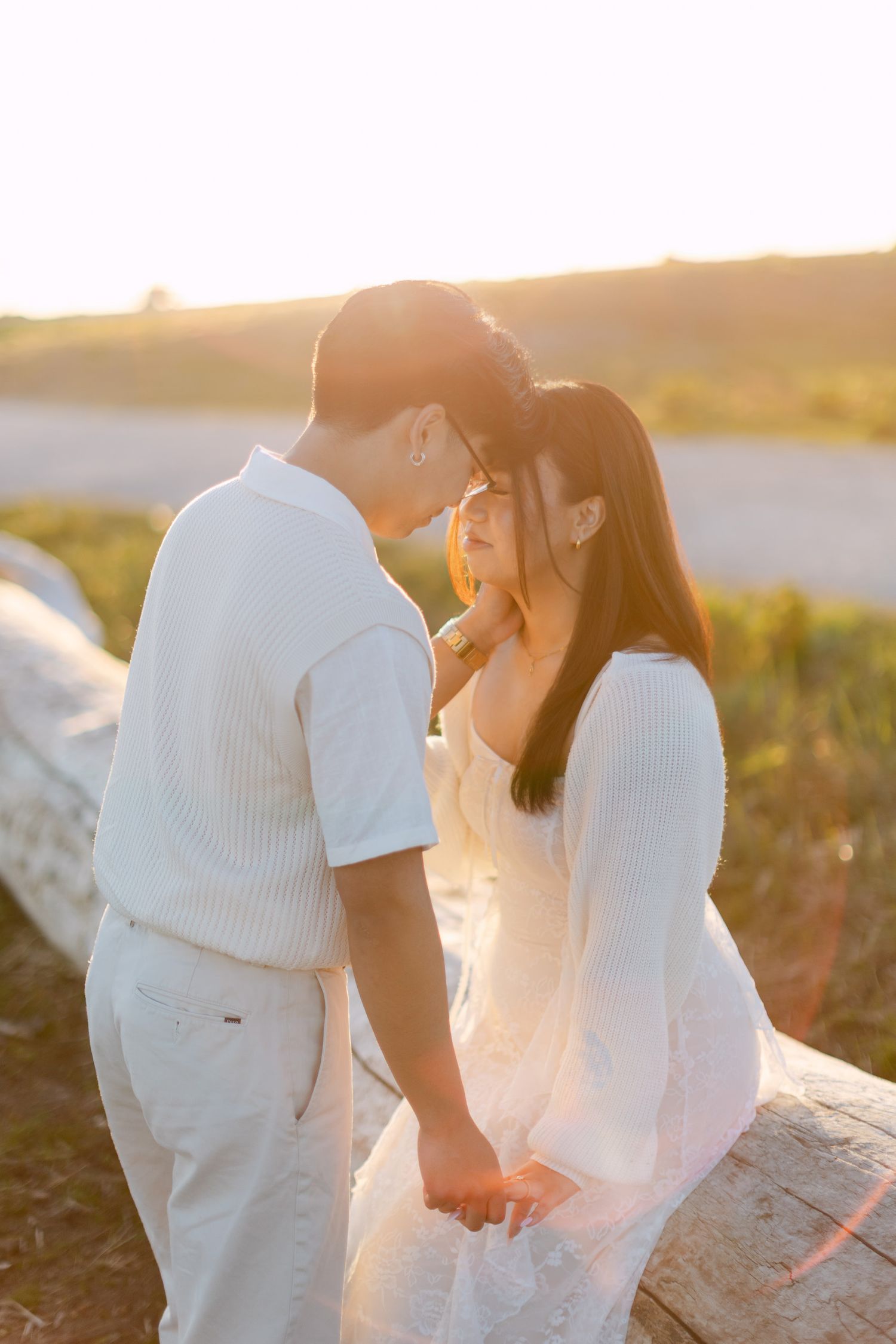 Romantic engagement photo of a couple embracing foreheads together on a fallen log in warm golden sunset light.
