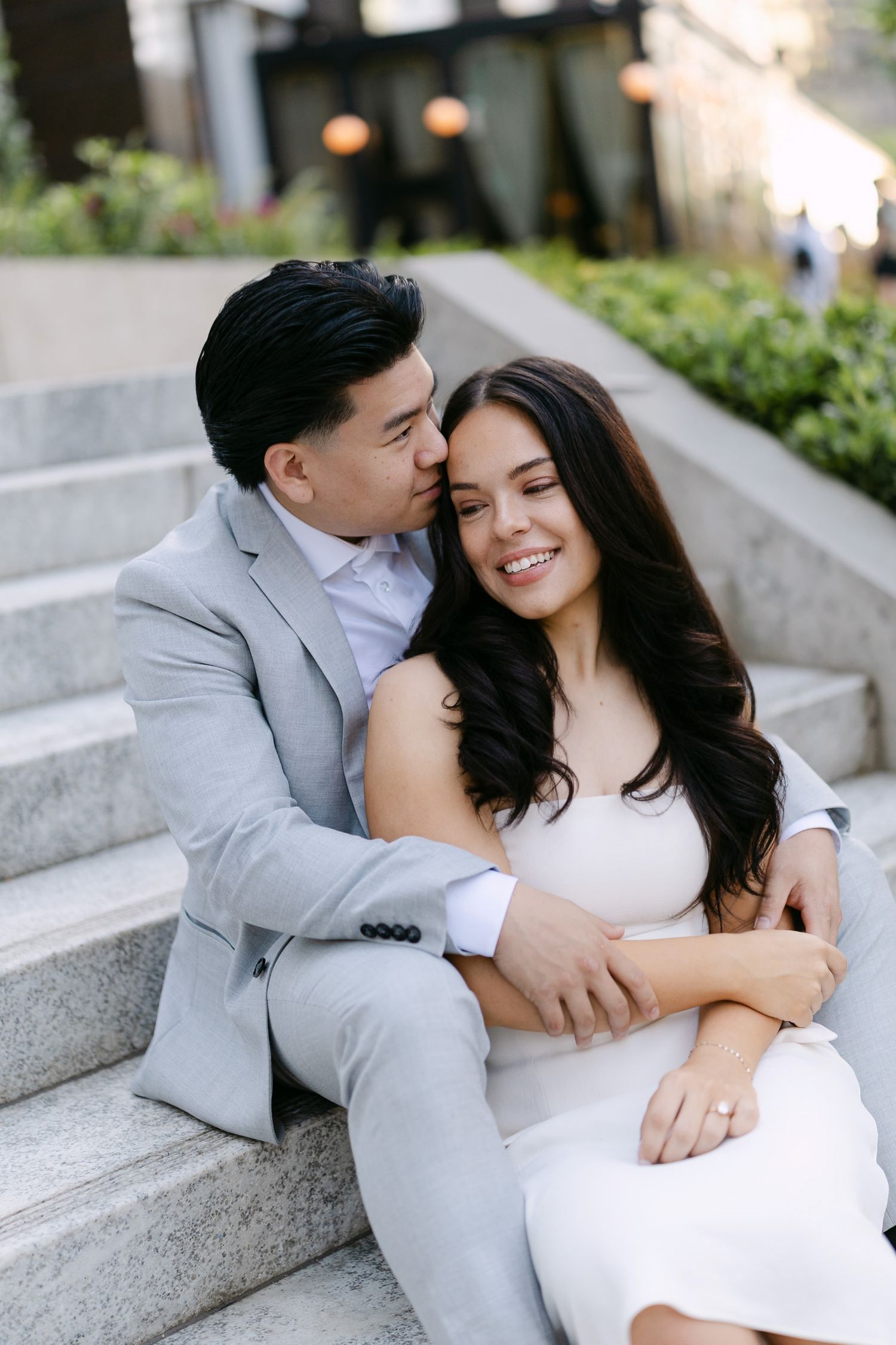 Smiling engaged couple sits close on stone steps, the man in a gray suit kissing her forehead as she wears a white dress.