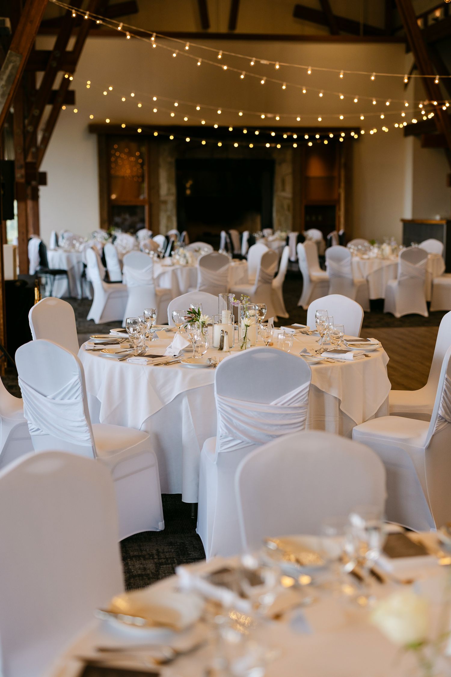 Elegant wedding reception hall with white chair covers, round tables, white linens, and warm string lights overhead.