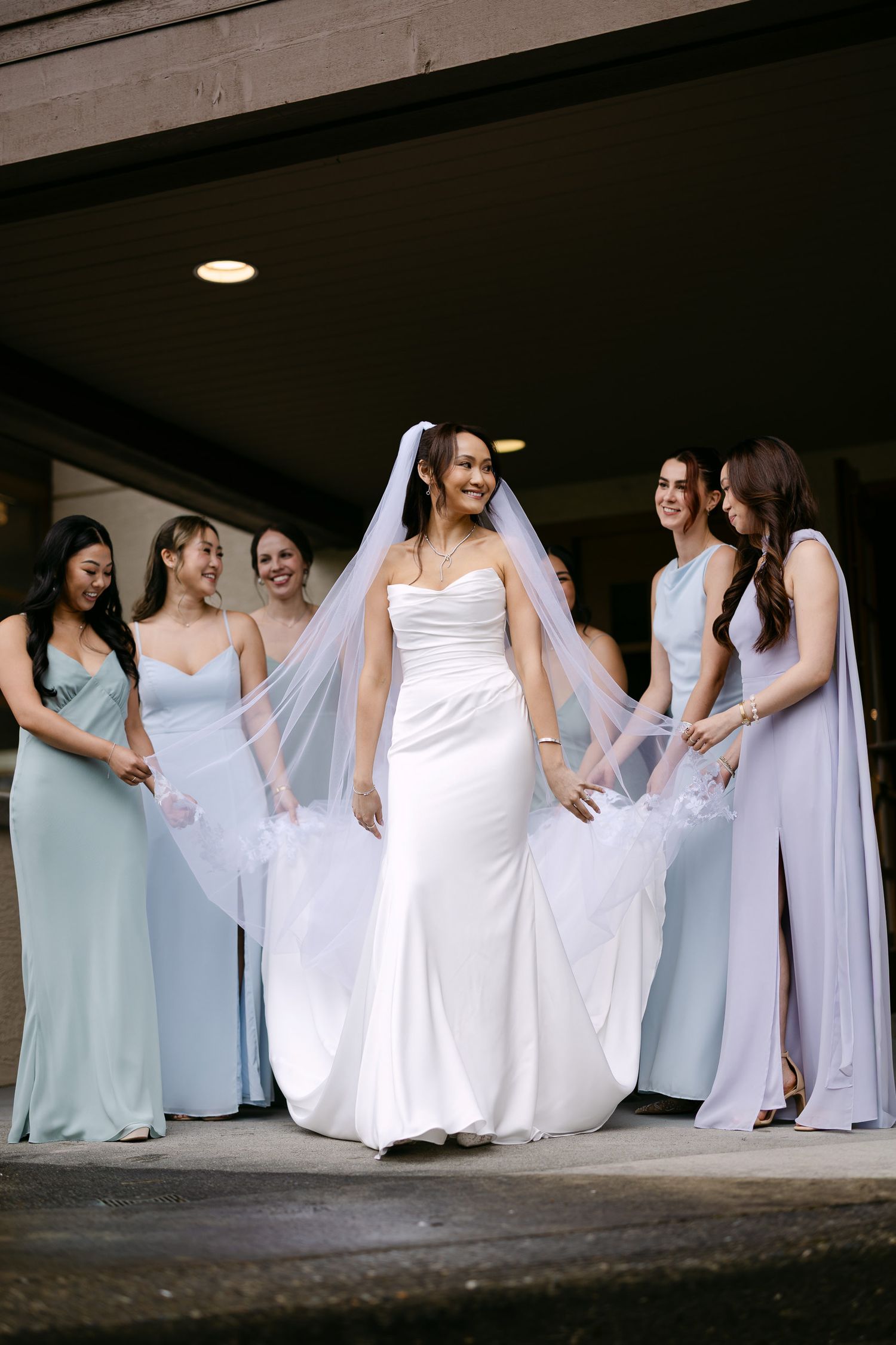 Bride in white gown and veil surrounded by smiling bridesmaids in light blue and green dresses at a wedding.