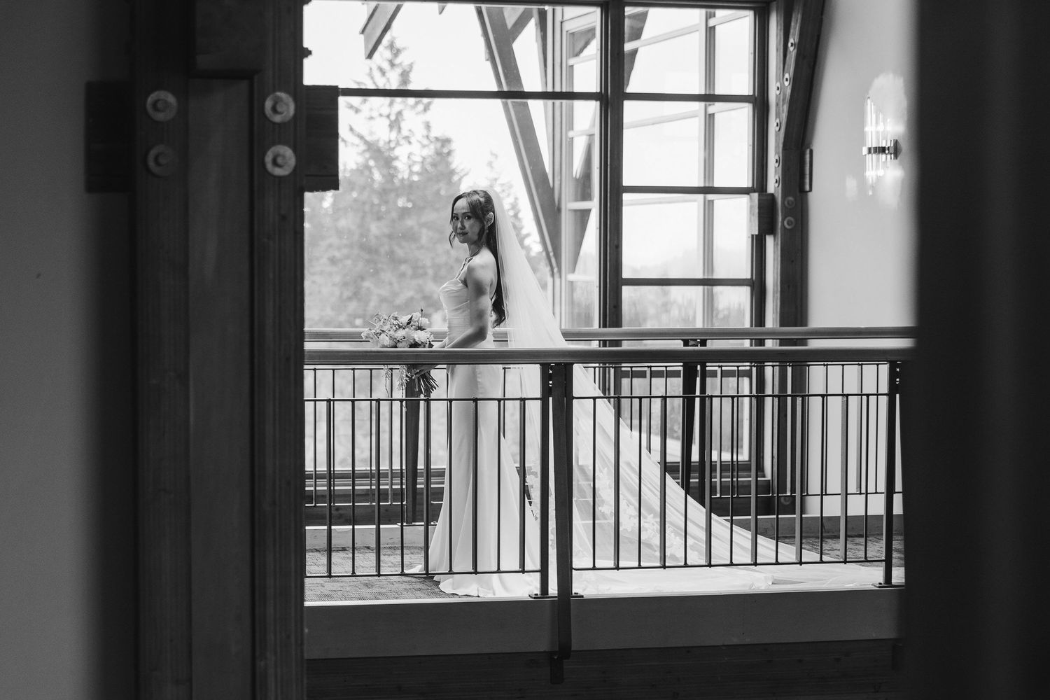 Black and white photo of a bride in wedding dress and veil standing on an indoor balcony gazing out large windows.