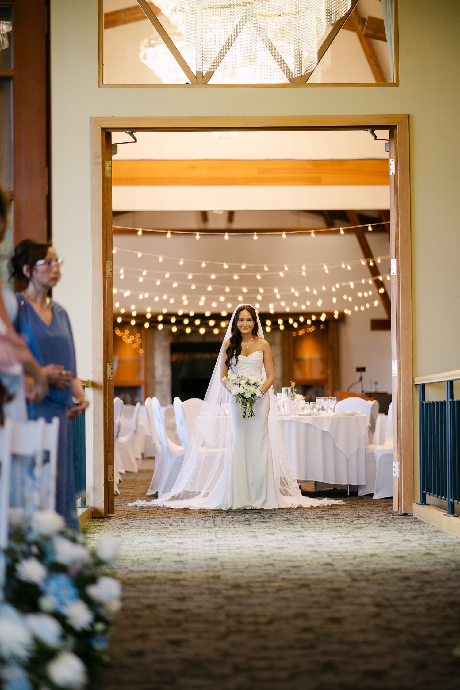 Smiling bride holding bouquet enters wedding reception hall adorned with string lights and white decor.