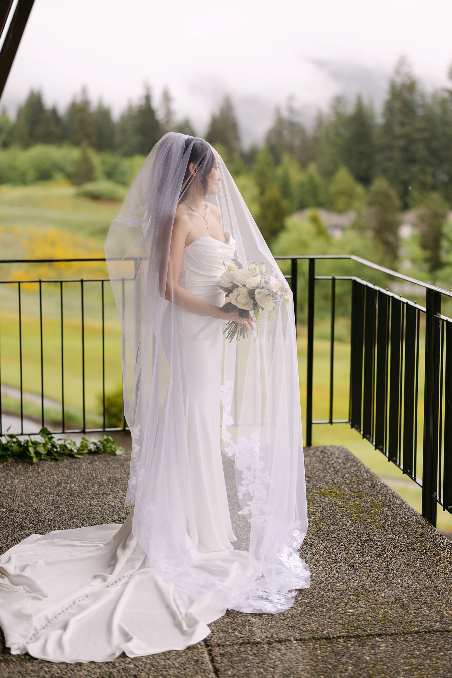 Bride in a strapless white gown with lace cathedral veil holds a bouquet on a misty outdoor balcony.