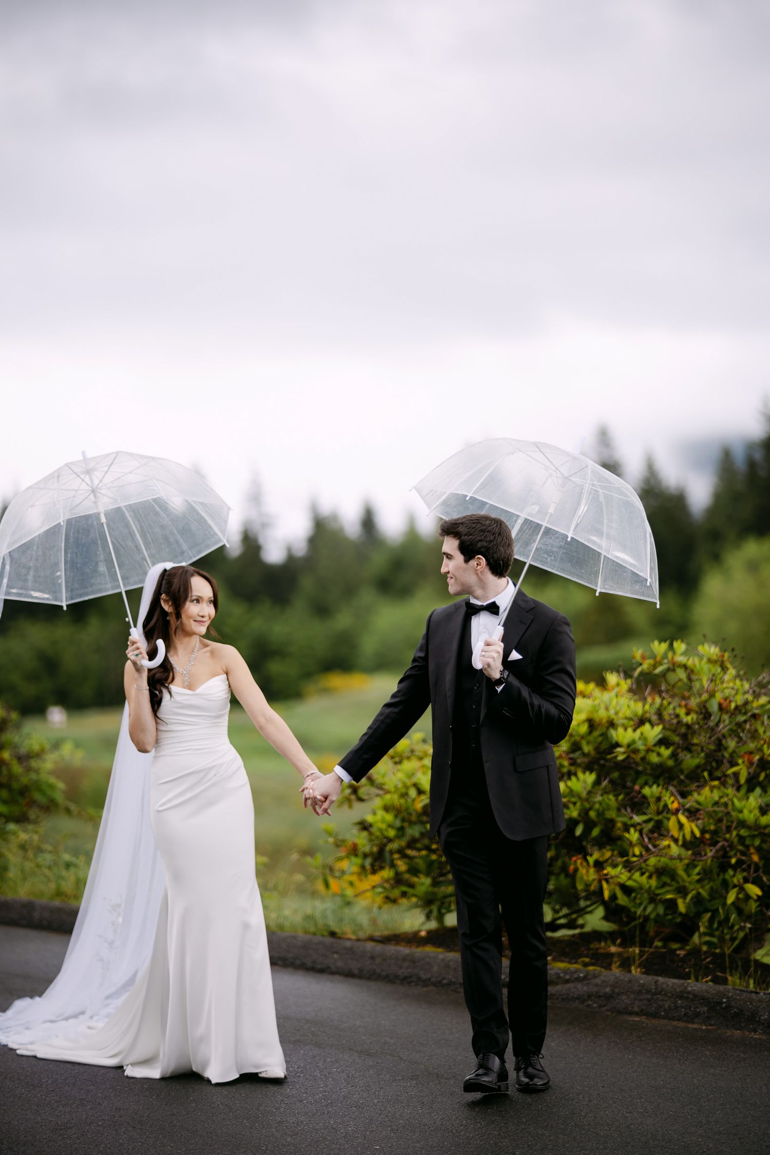Bride and groom hold clear umbrellas and walk hand-in-hand on rainy day with lush greenery behind them.