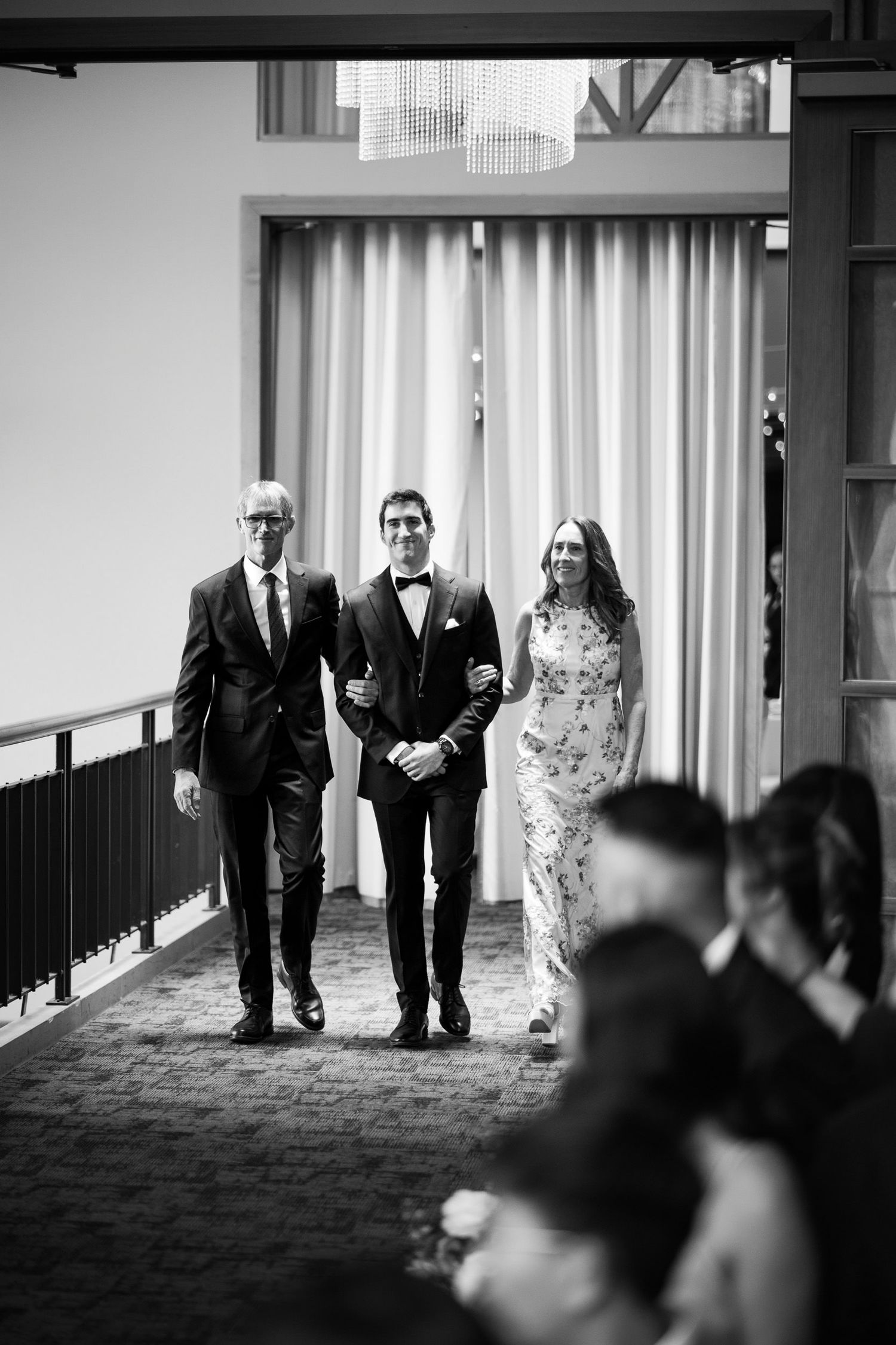 Groom walks down the aisle escorted by parents in black and white wedding ceremony photo.