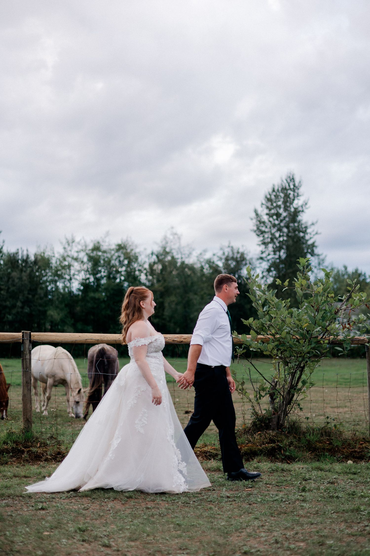 A wedding couple walks through a rustic field with trees and cloudy skies in the background.