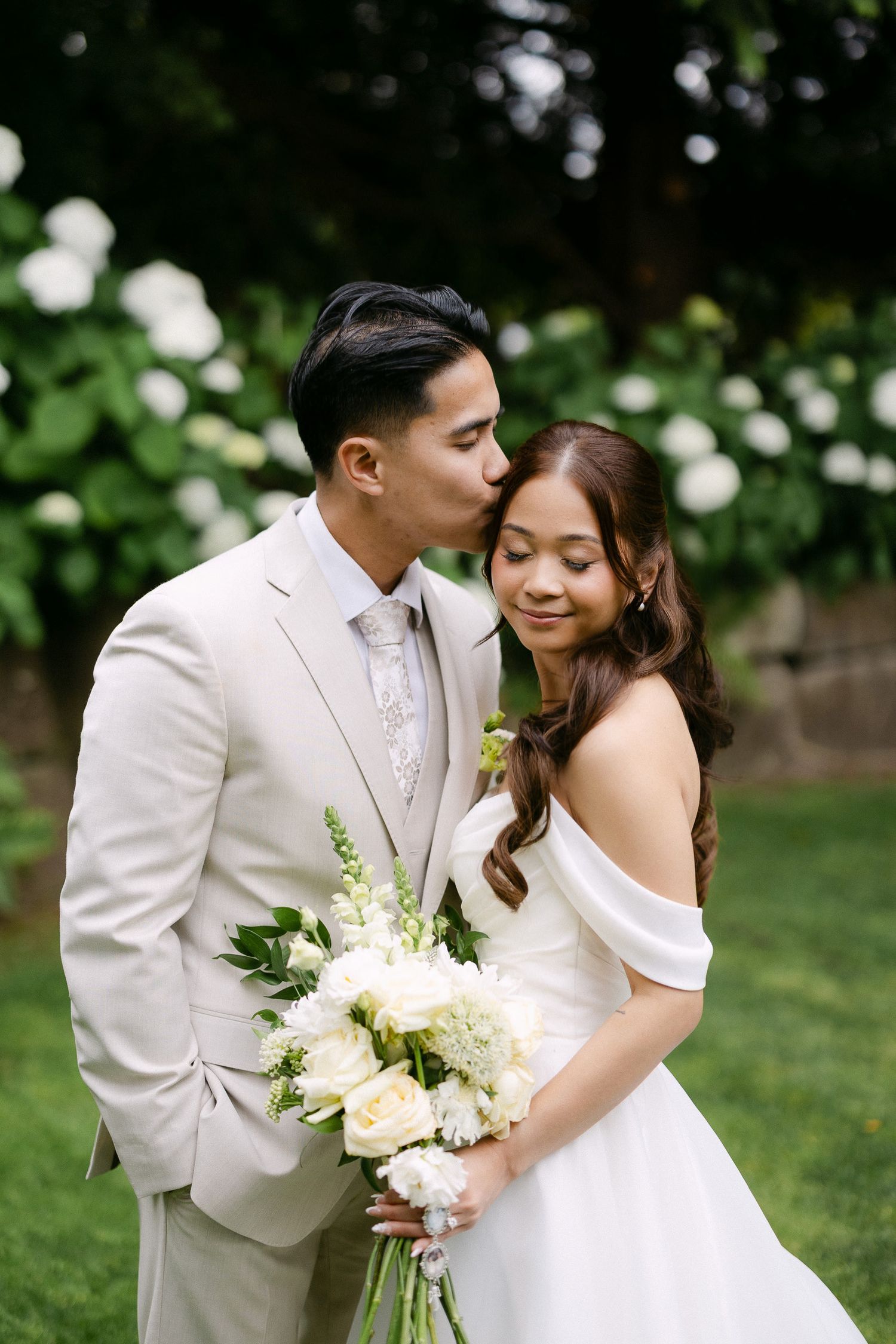 Groom in beige suit kisses bride wearing off-shoulder white gown holding white floral bouquet outdoors.