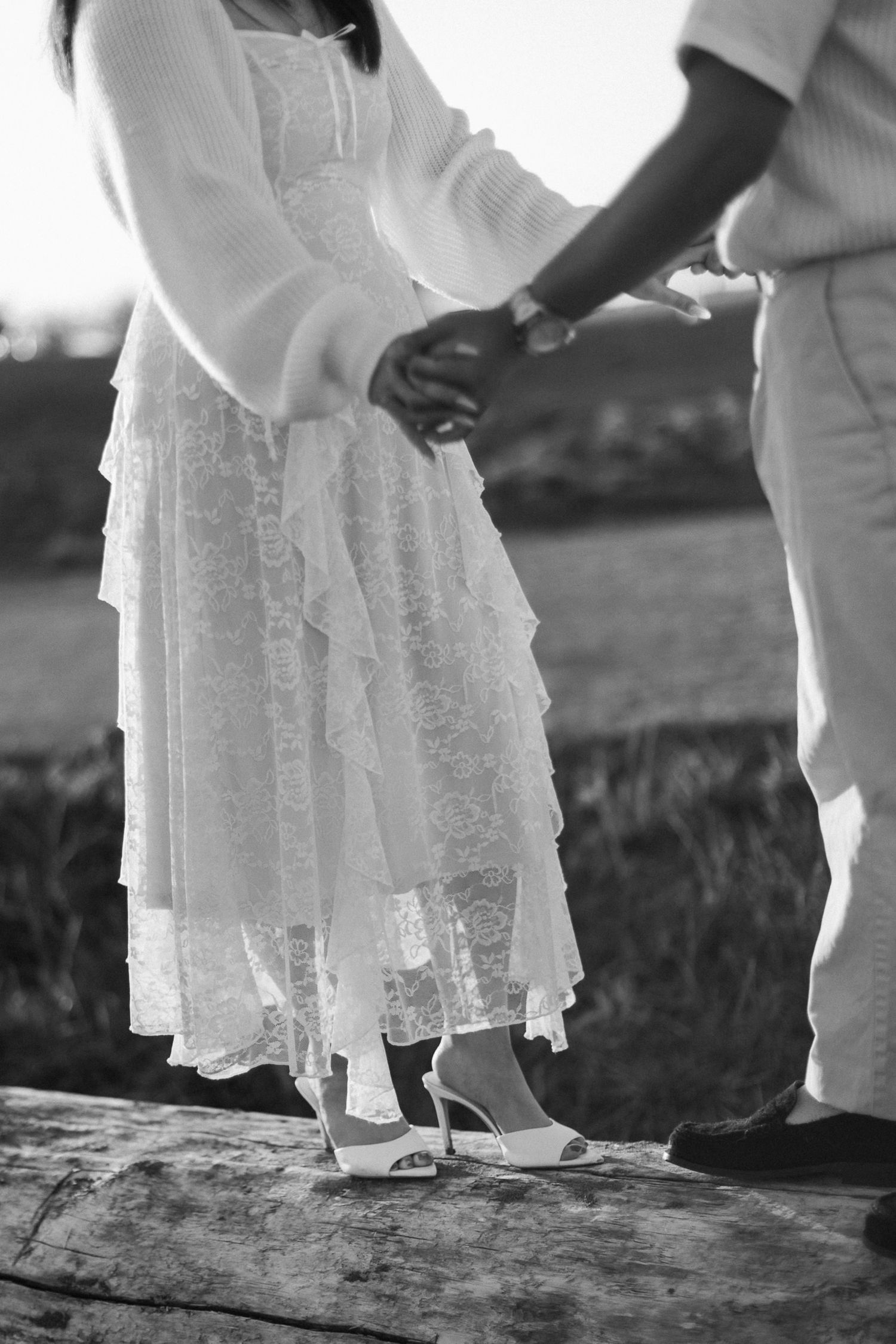 Black and white image of a couple holding hands, the woman in a ruffled lace dress and white heels standing on a log.