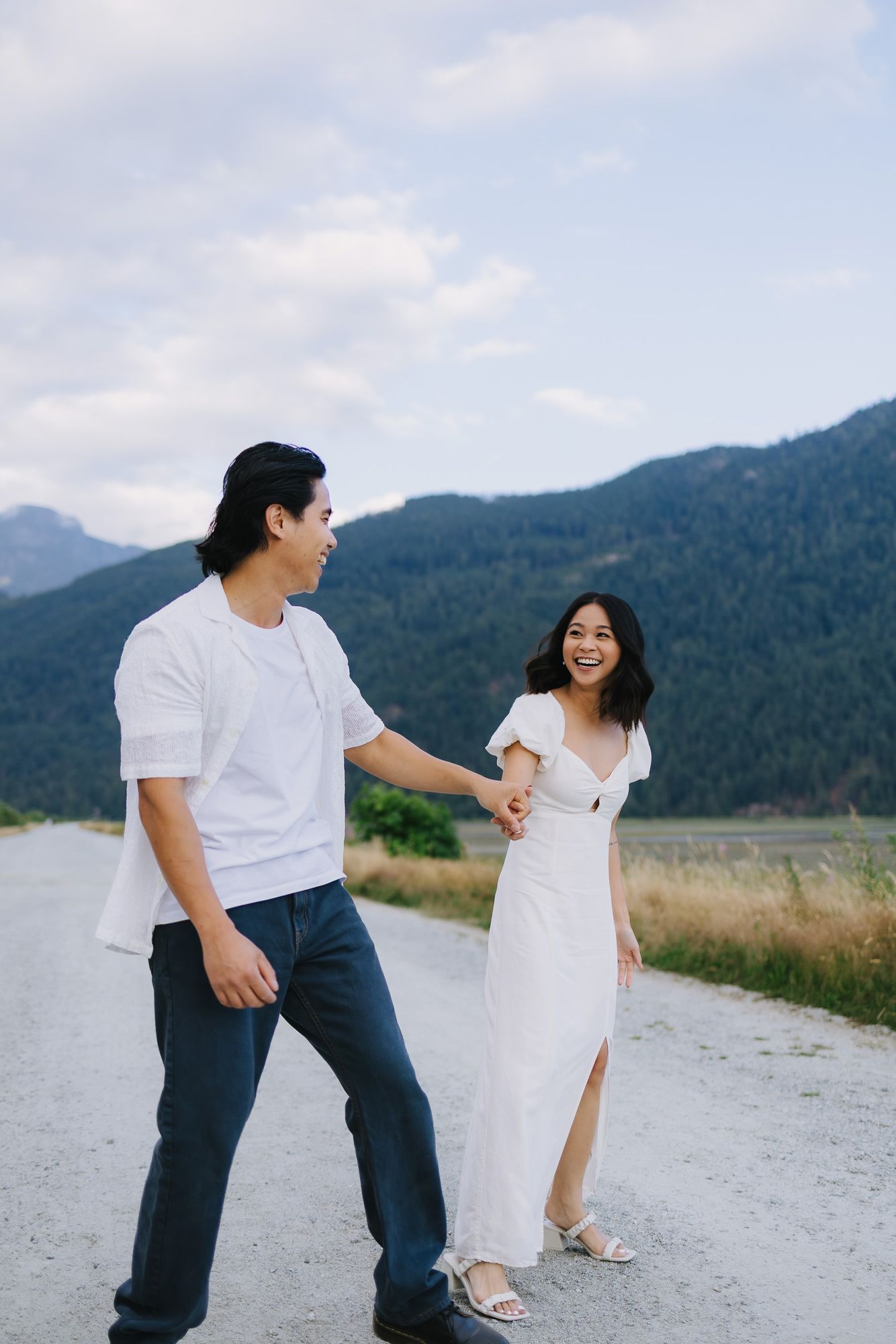 Laughing couple in white outfits hold hands and stroll along a gravel road with forested mountains and cloudy sky behind.