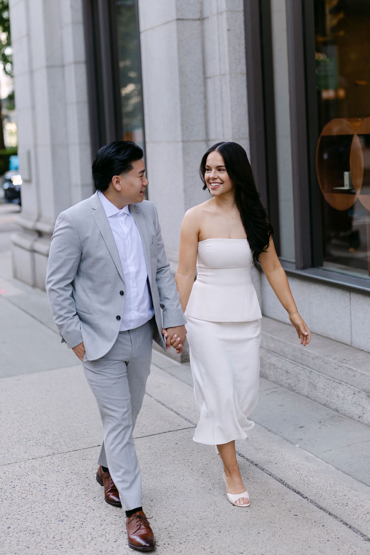 Smiling couple in a gray suit and white dress walk hand in hand along a city sidewalk past a stone building.