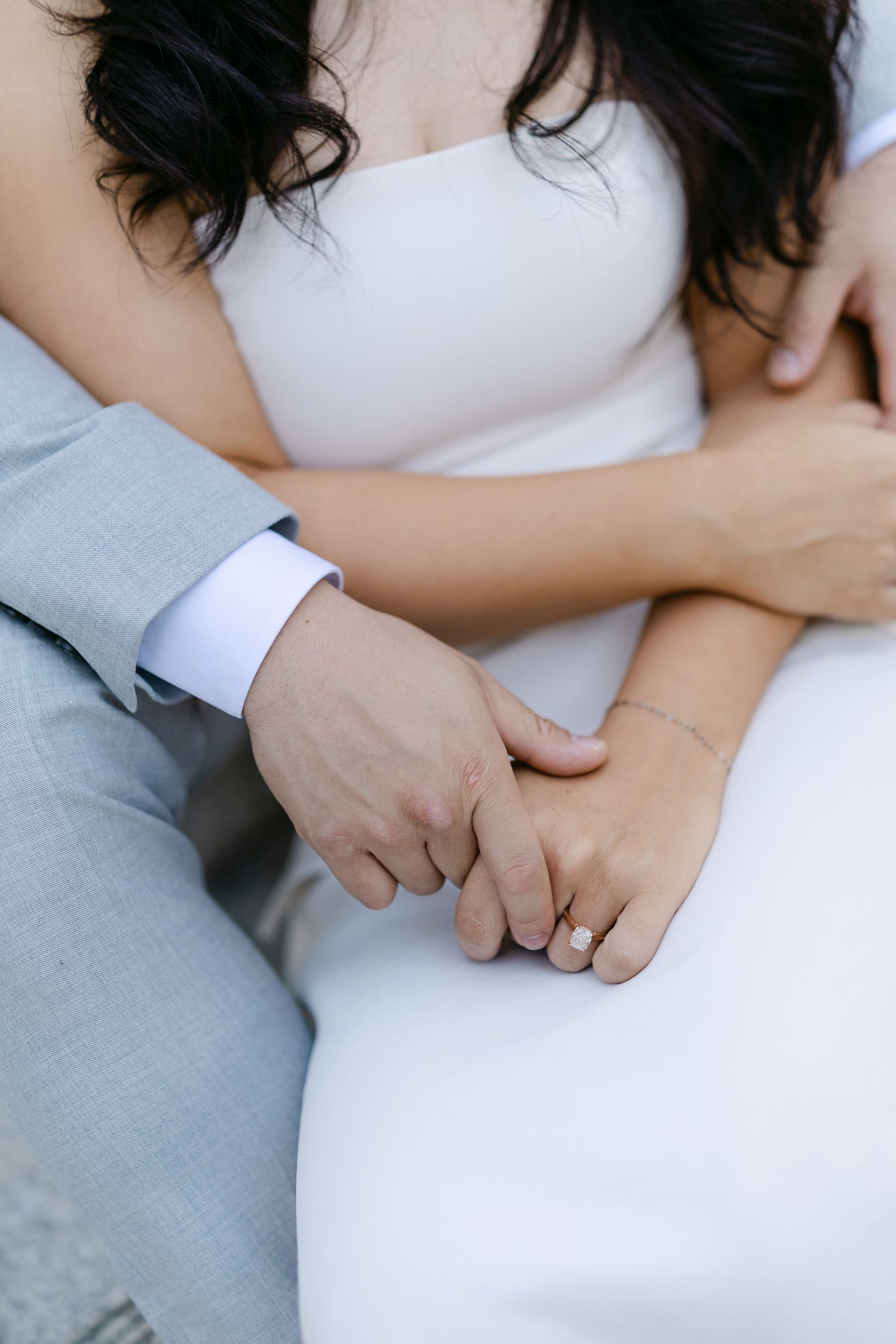 Close-up of a couple's intertwined hands, the bride in a white dress wearing a diamond ring and bracelet, the groom in a gray suit.