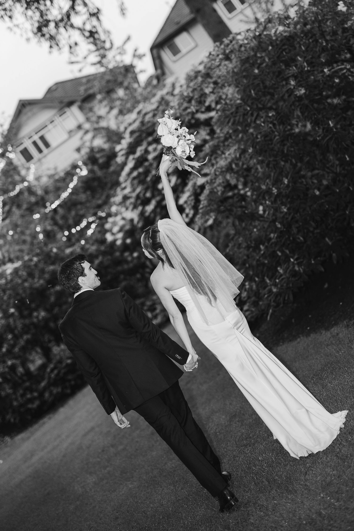 Black and white photo of bride and groom walking hand in hand, bride raising bouquet joyfully.