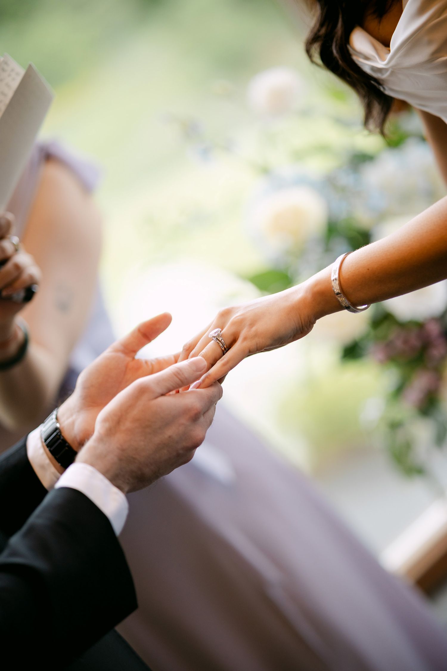 Groom placing a wedding ring on brides finger during ceremony, with flowers in the background.