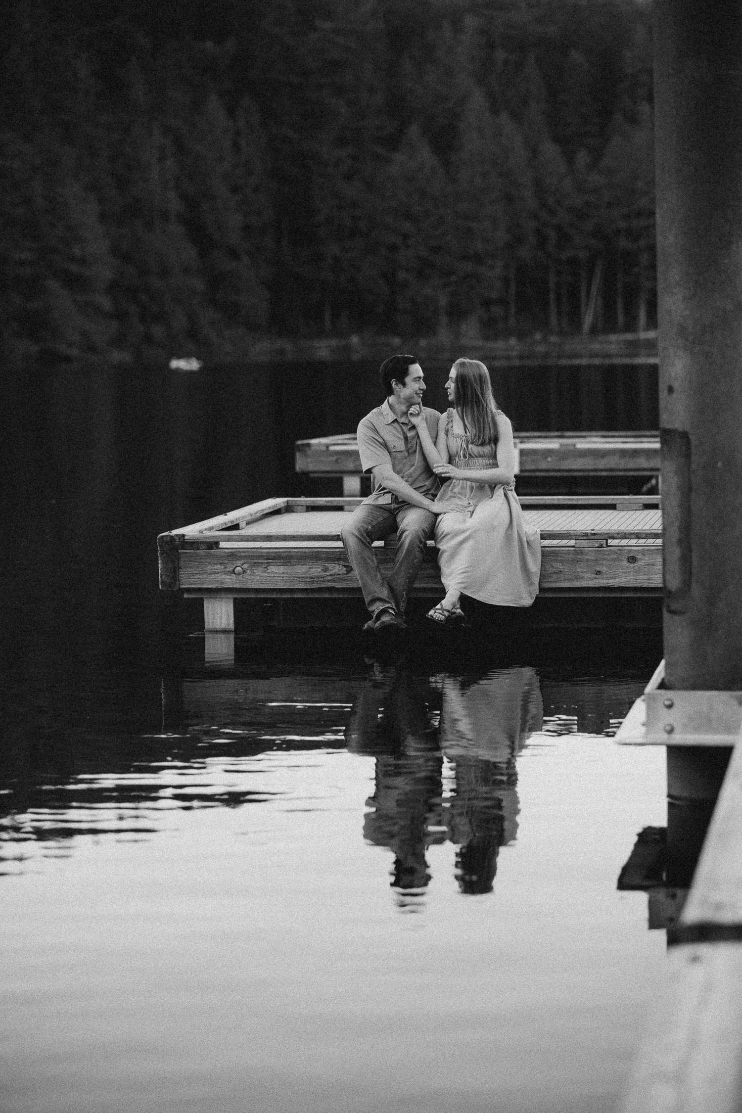 A couple sits together on a wooden dock by a calm lake in black and white.