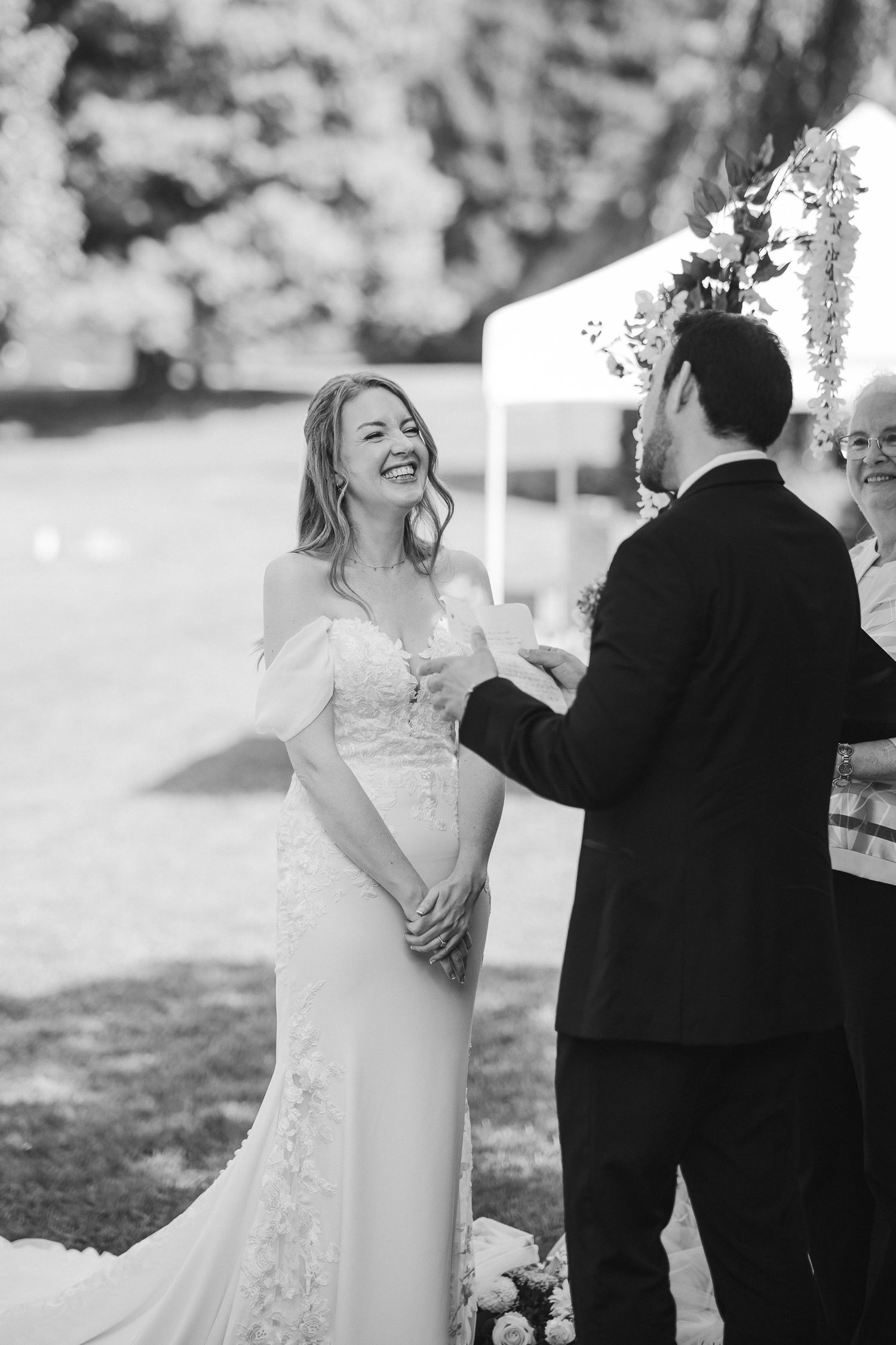 Black and white photo of a joyful wedding ceremony moment between a bride in a fitted lace gown and groom in a dark suit.