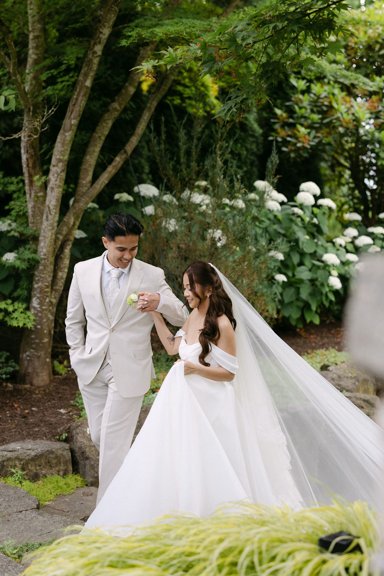Smiling bride and groom walk through a green garden, her white ballgown veil flowing behind them.