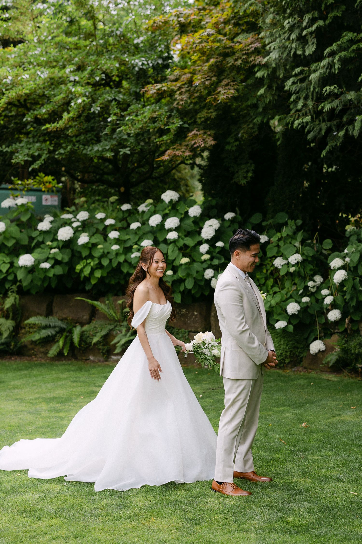 Bride in off-shoulder ball gown smiles at groom in tan suit during first look, surrounded by white hydrangeas.