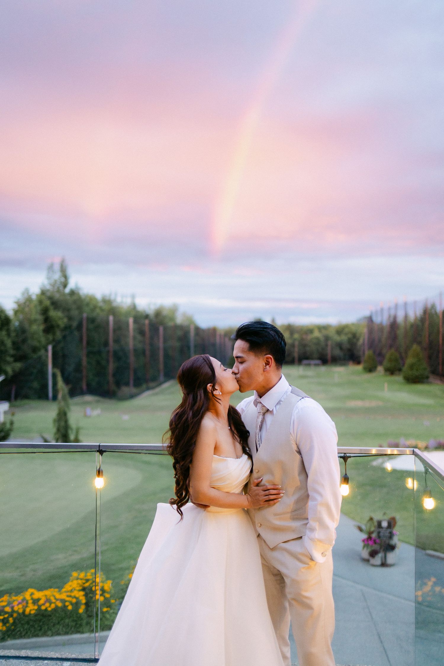 Bride and groom share a kiss on a balcony at sunset with a rainbow in the sky behind them.