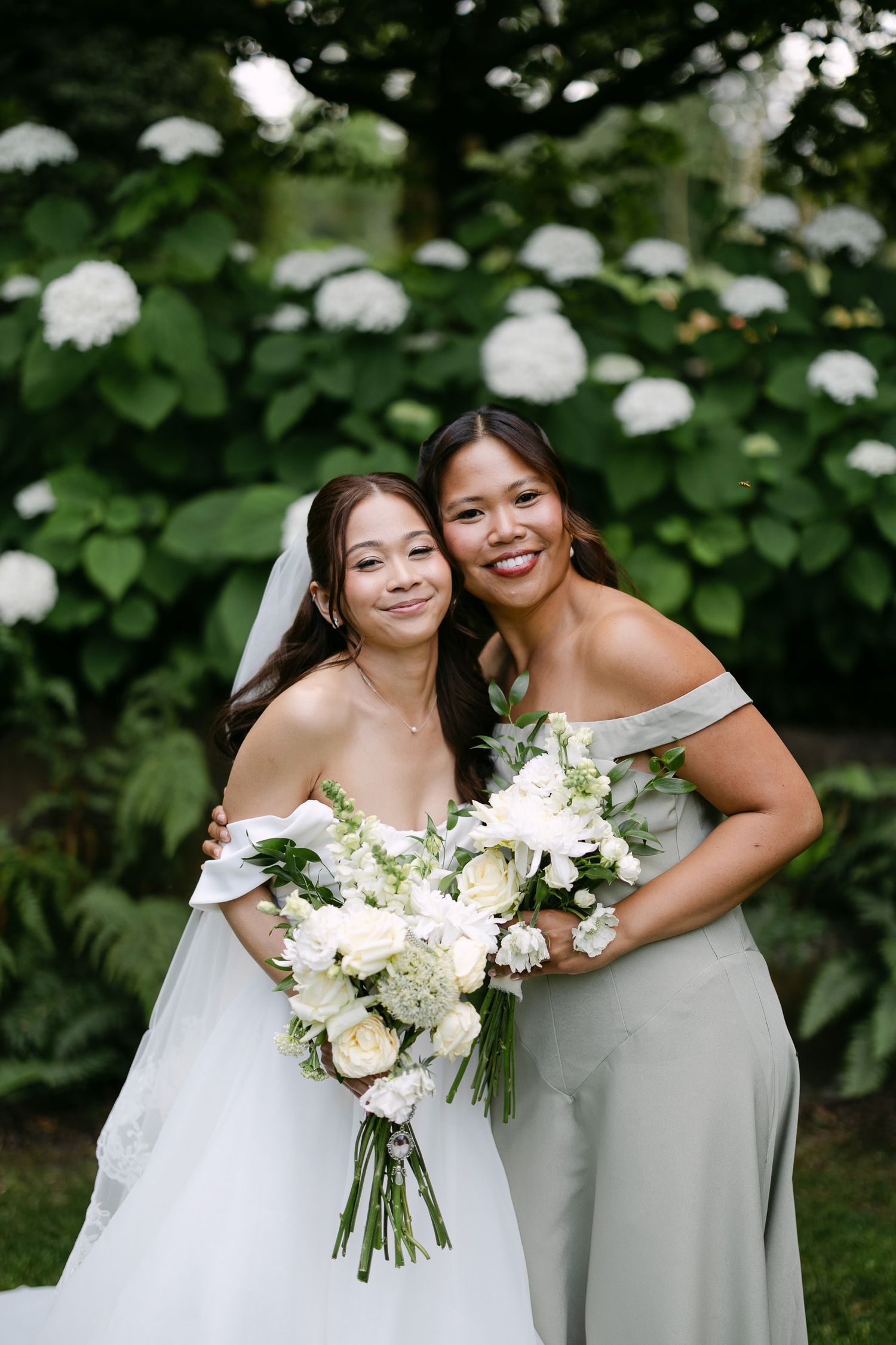 Bride and bridesmaid smile together holding white floral bouquets in front of hydrangea bushes.