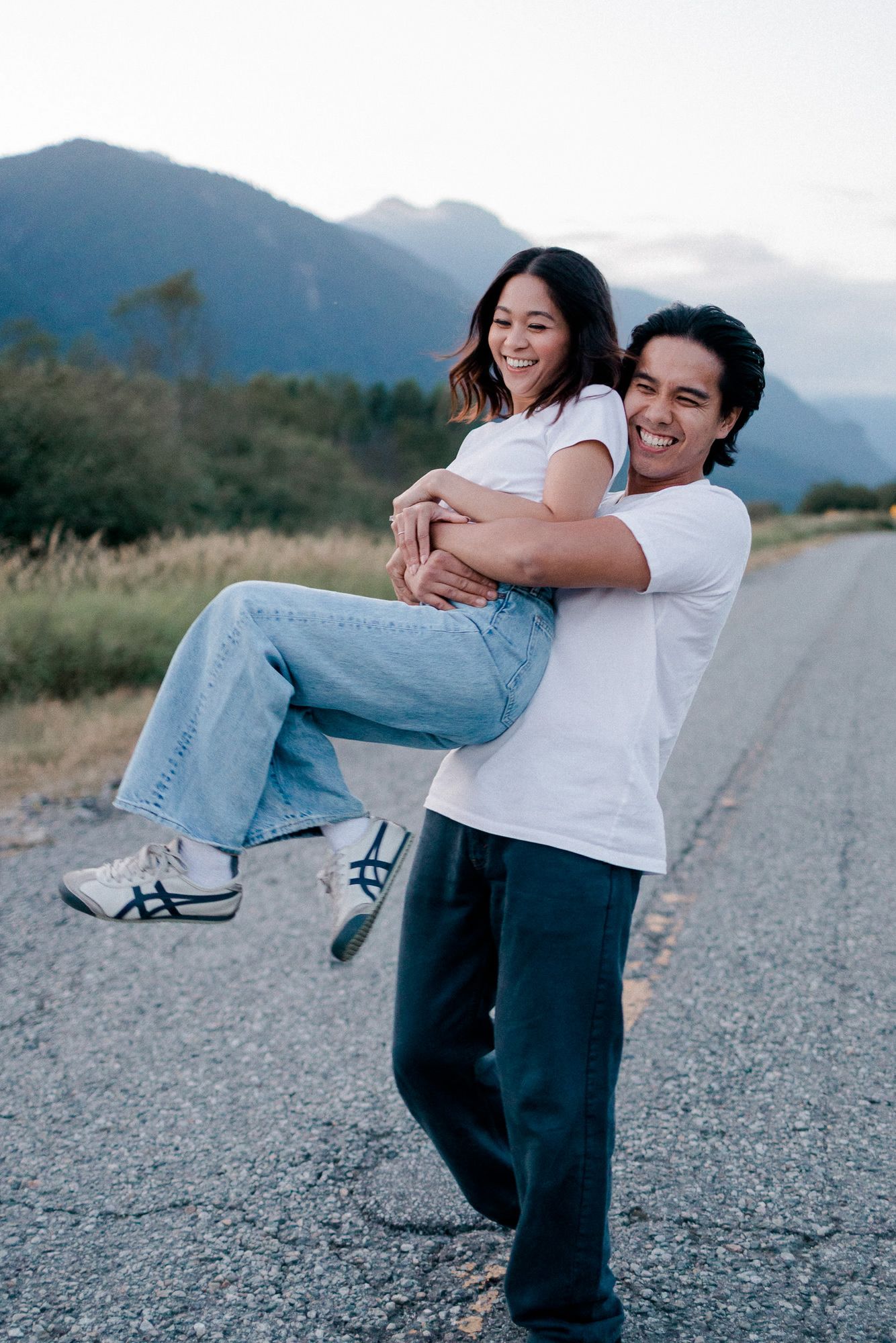 A smiling man lifts a laughing woman in his arms on a quiet paved road with mountains in the background.