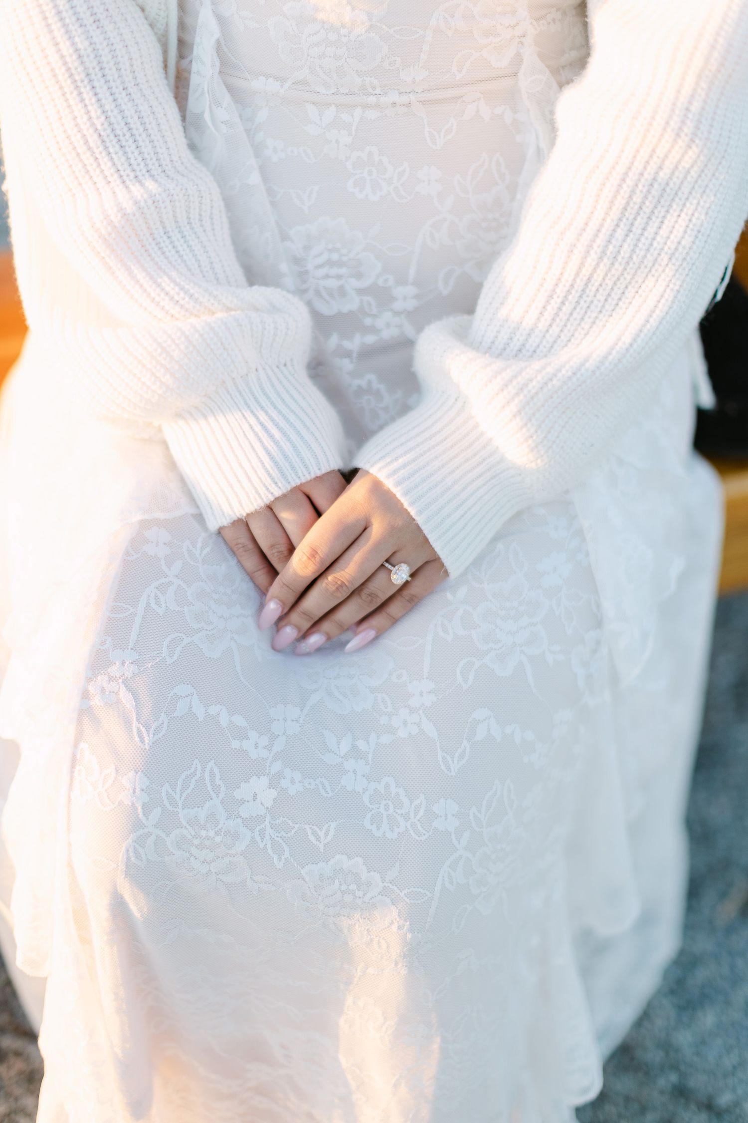 Close-up of a bride-to-be's hands wearing a diamond engagement ring, resting on a white lace dress and cream cardigan.
