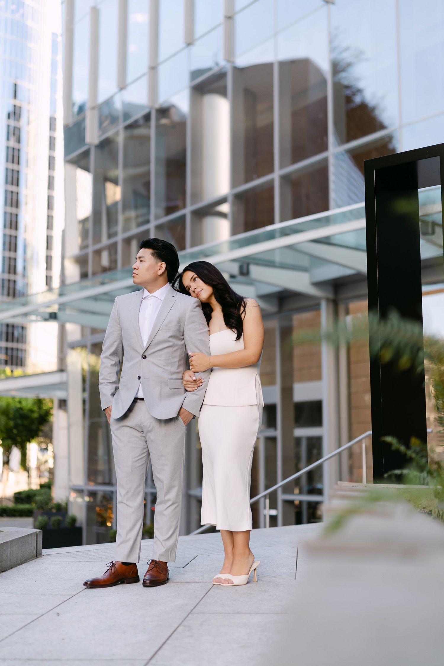 Couple posing outdoors in front of a modern glass building, the woman in a white dress leaning on a man in a light gray suit.