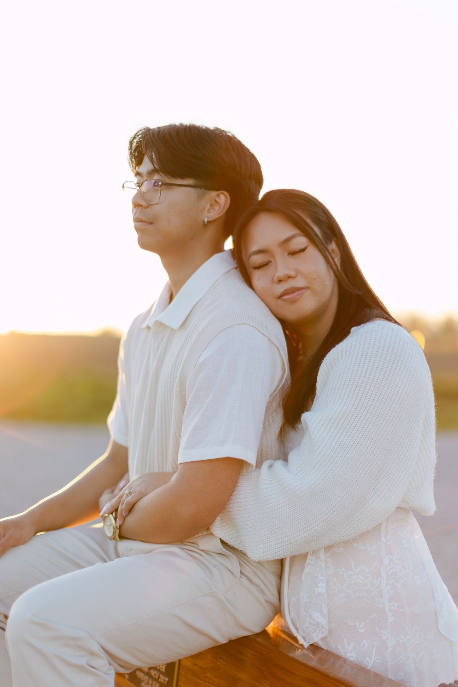 Woman rests her head on her partner's shoulder as they sit together on a bench in warm sunset light.